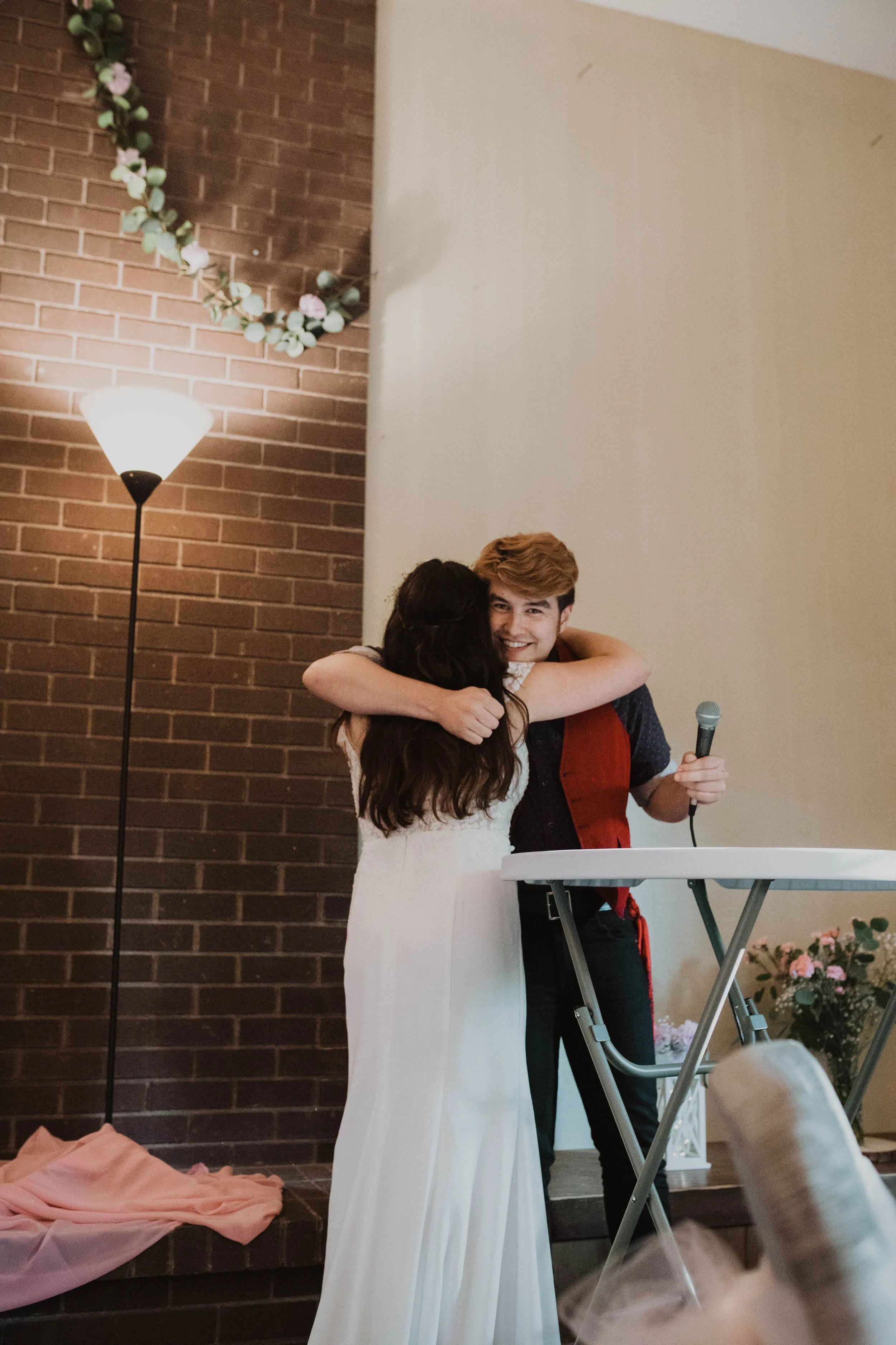 A young man and woman hugging at a celebration, with the man holding a microphone, in a decorated indoor setting with a brick wall, flowers, and a lamp. Seattle, WA wedding photography.