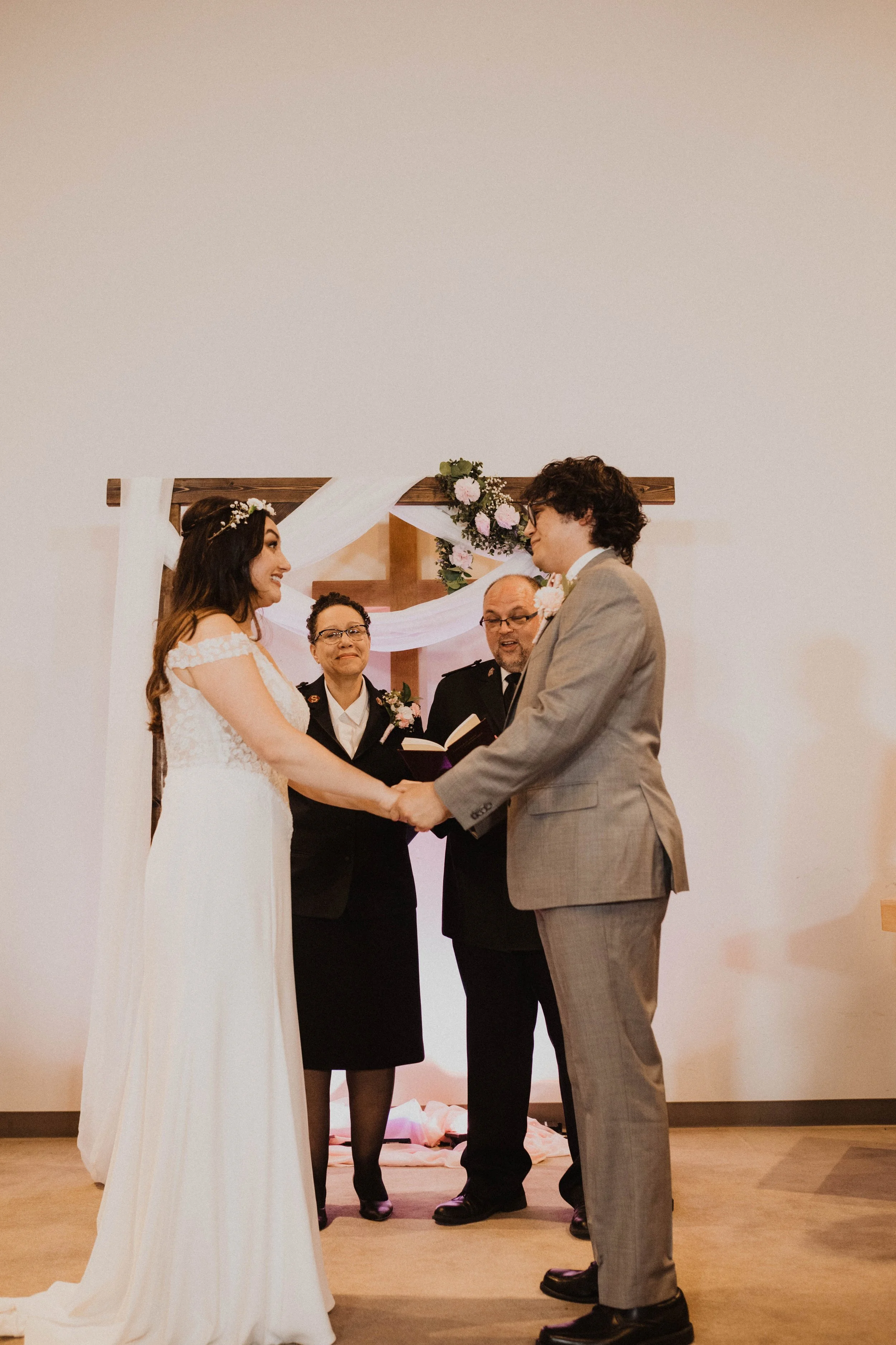 A bride and groom holding hands during a wedding ceremony, standing in front of officiants and a decorated wooden cross with flowers and white fabric. Seattle, WA wedding photography.