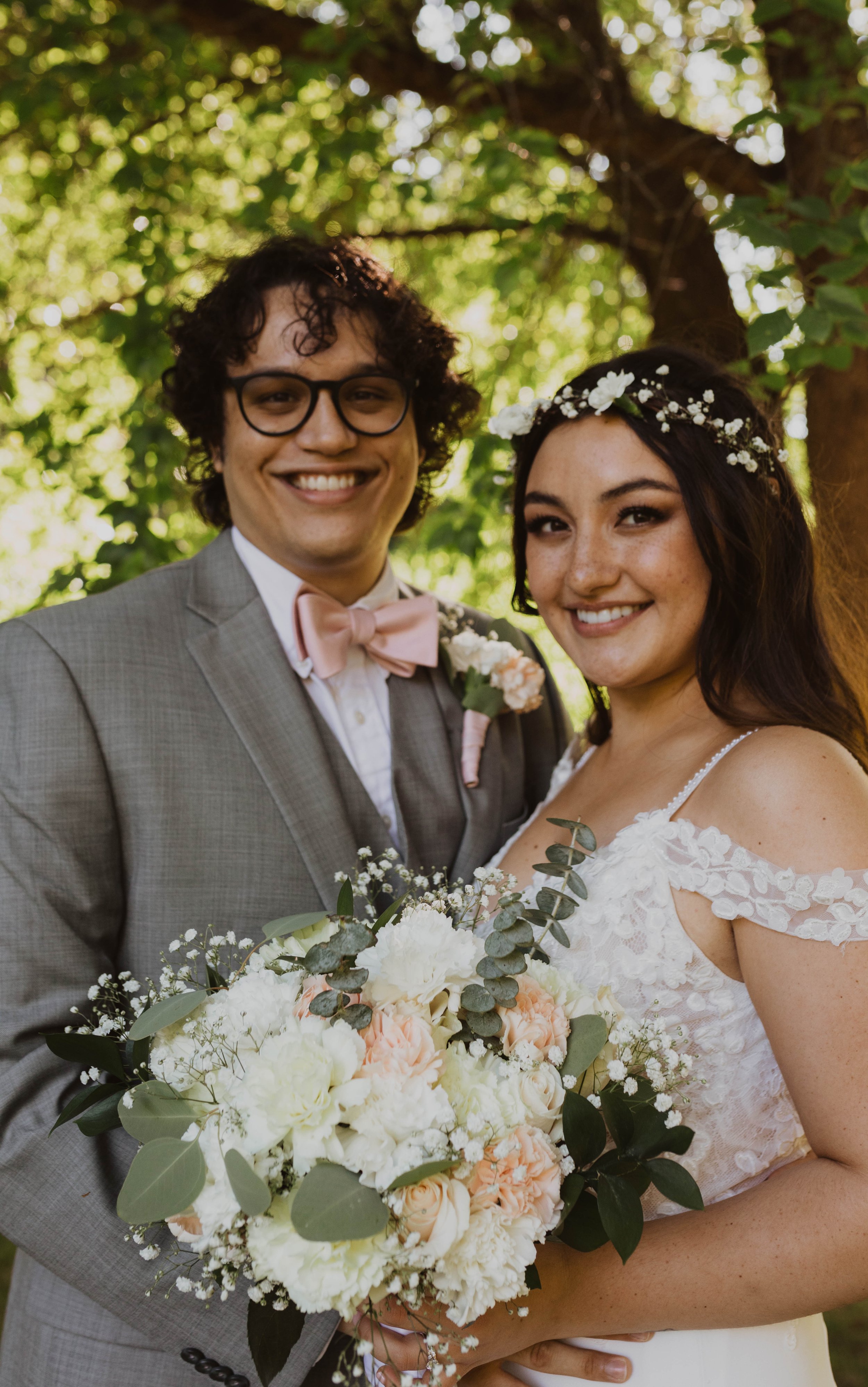 A newlywed couple standing outdoors under a tree, smiling at the camera. The groom is wearing glasses, a gray suit, a pink bowtie, and a boutonniere. The bride is wearing a white lace wedding gown with off-the-shoulder sleeves and a floral headband. 