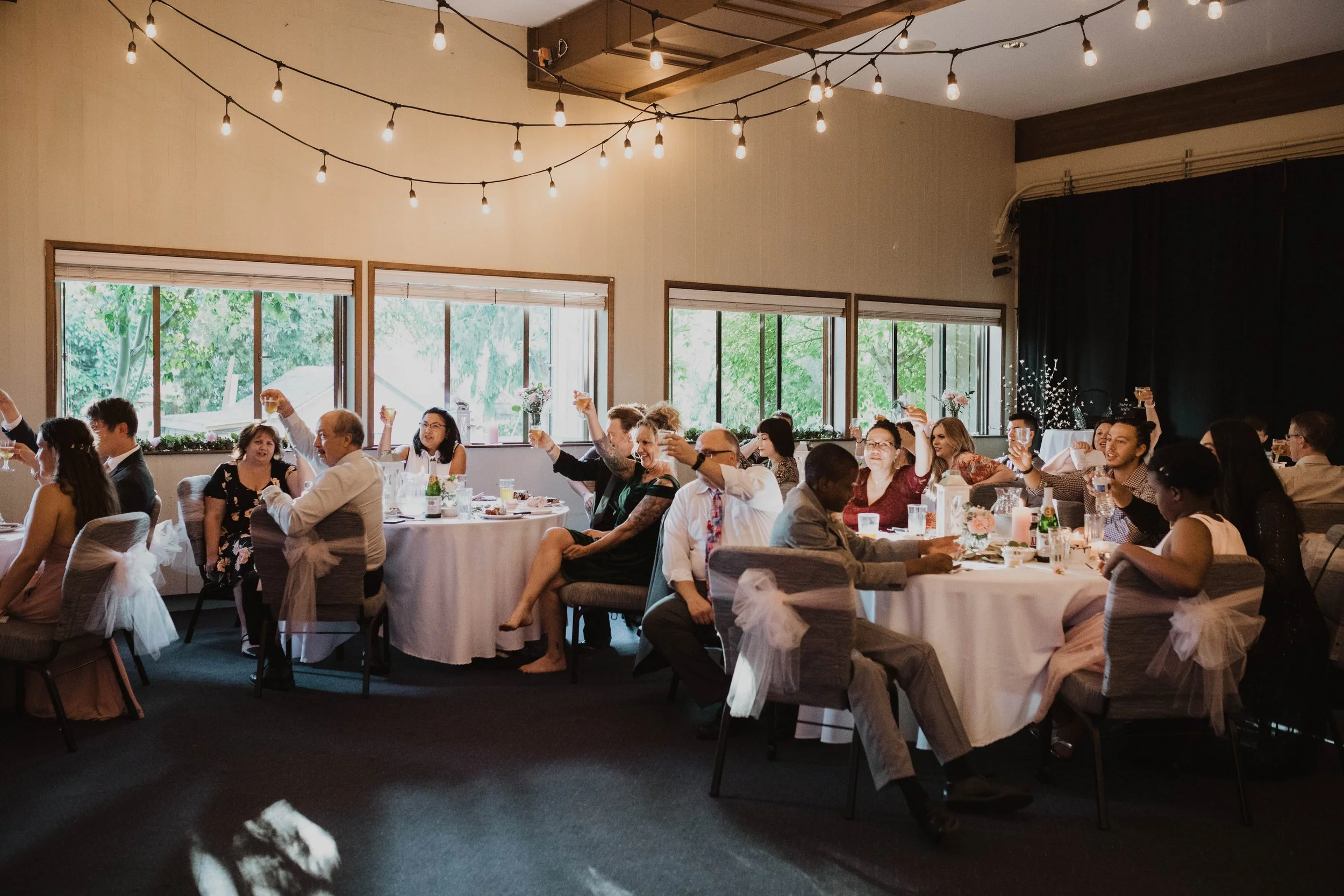 Guests at a wedding reception sitting at round tables, raising glasses in toast, decorated with white tablecloths, tulle bows on chairs, and surrounded by windows showing greenery outside. Seattle, WA wedding photography.