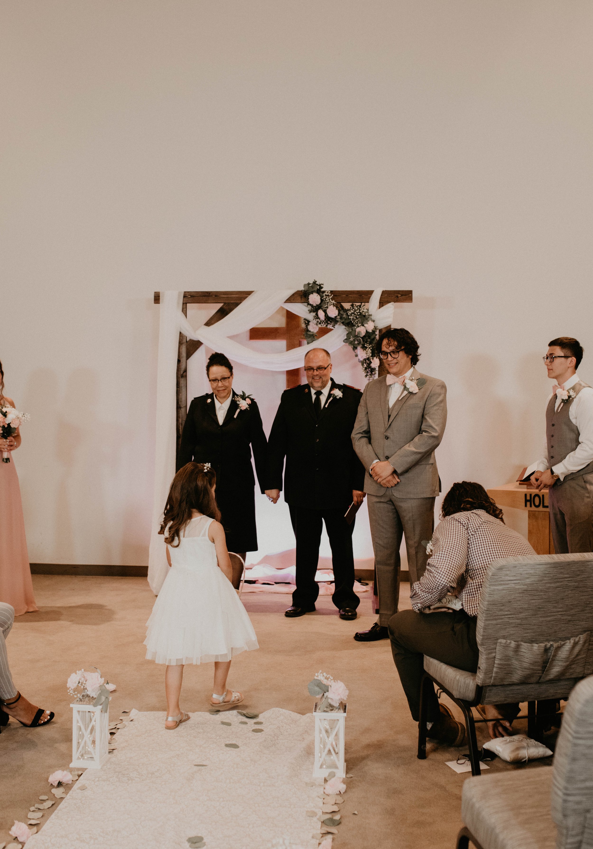 A young girl in a white dress walking down a wedding aisle in front of an officiant and wedding party, with a decorated wooden arch in the background. Seattle, WA wedding photography.