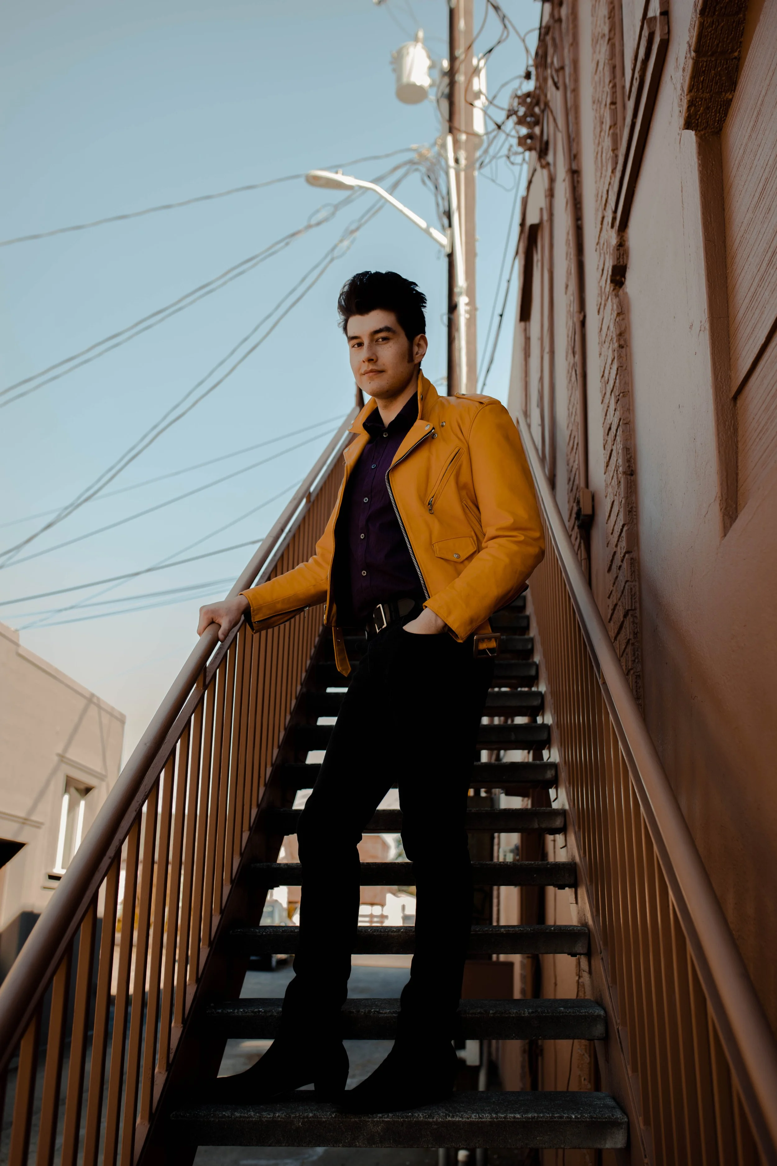 A young man with dark hair, wearing a yellow jacket and black pants, standing on outdoor metal stairs against a tan building and blue sky. Seattle professional head shot photography
