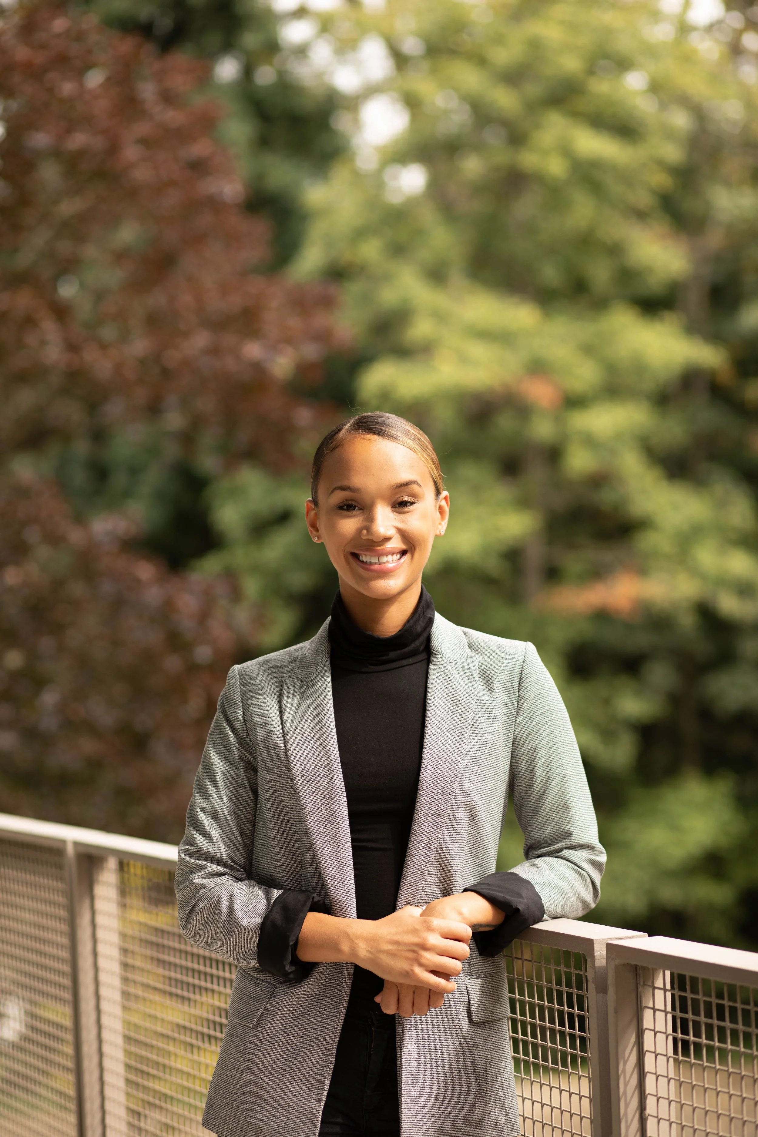 A professional woman with a friendly smile, standing outdoors on a balcony or terrace, with green trees in the background. Seattle professional head shot photography