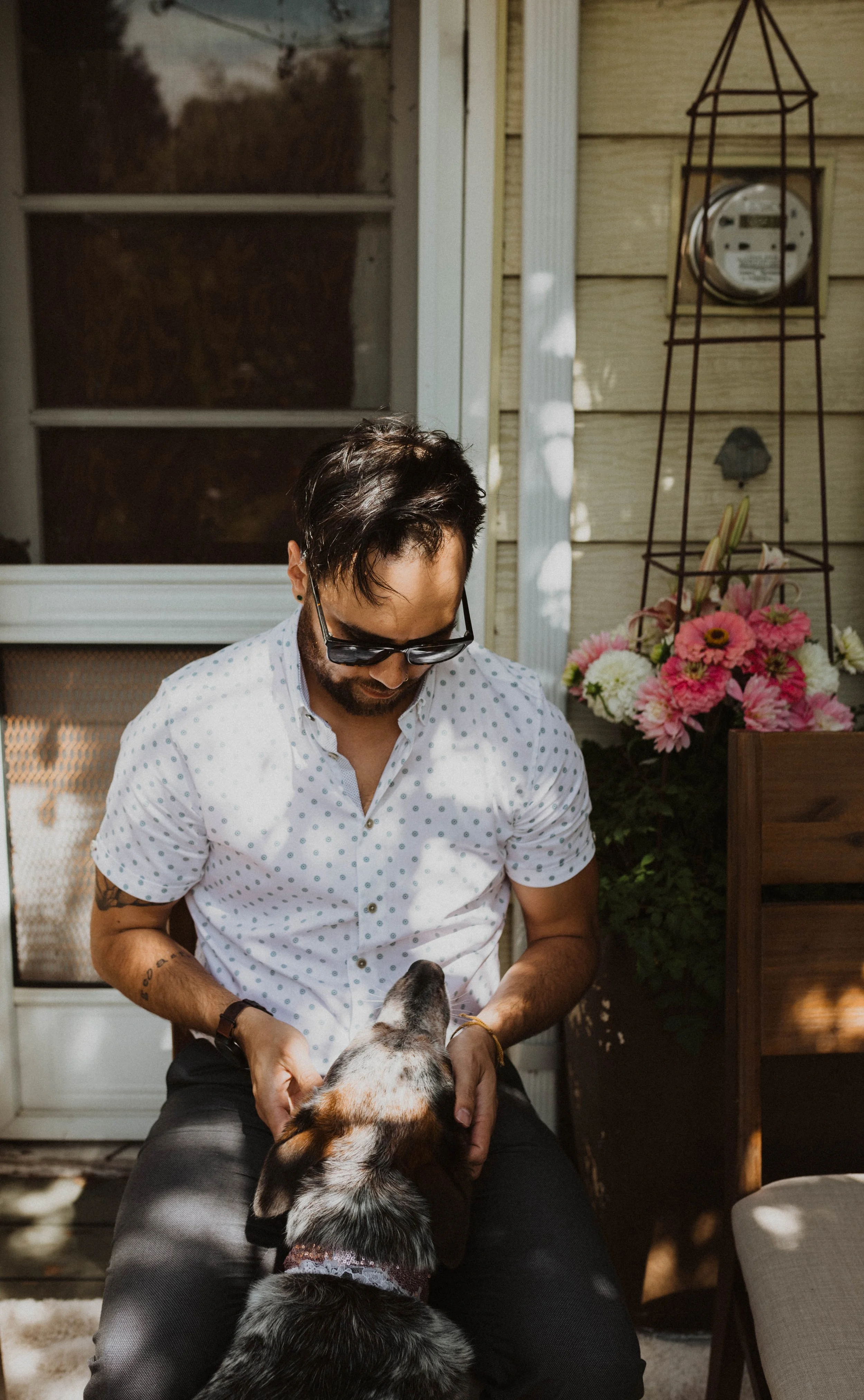A man wearing sunglasses and a white polka dot shirt sits on a porch, gently holding a dog’s face as the dog looks up at him. Seattle, WA wedding photography.