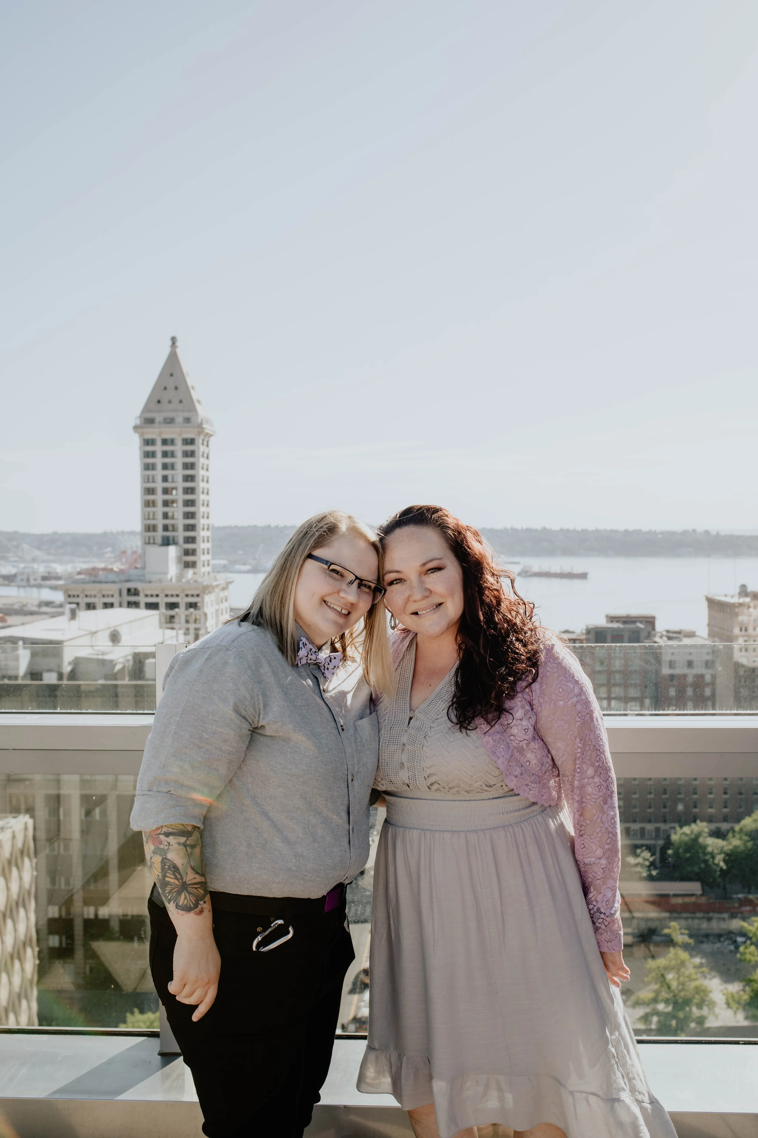 Two women smiling and hugging on a balcony with a city skyline and waterfront in the background. Seattle Municipal Courthouse wedding photography.