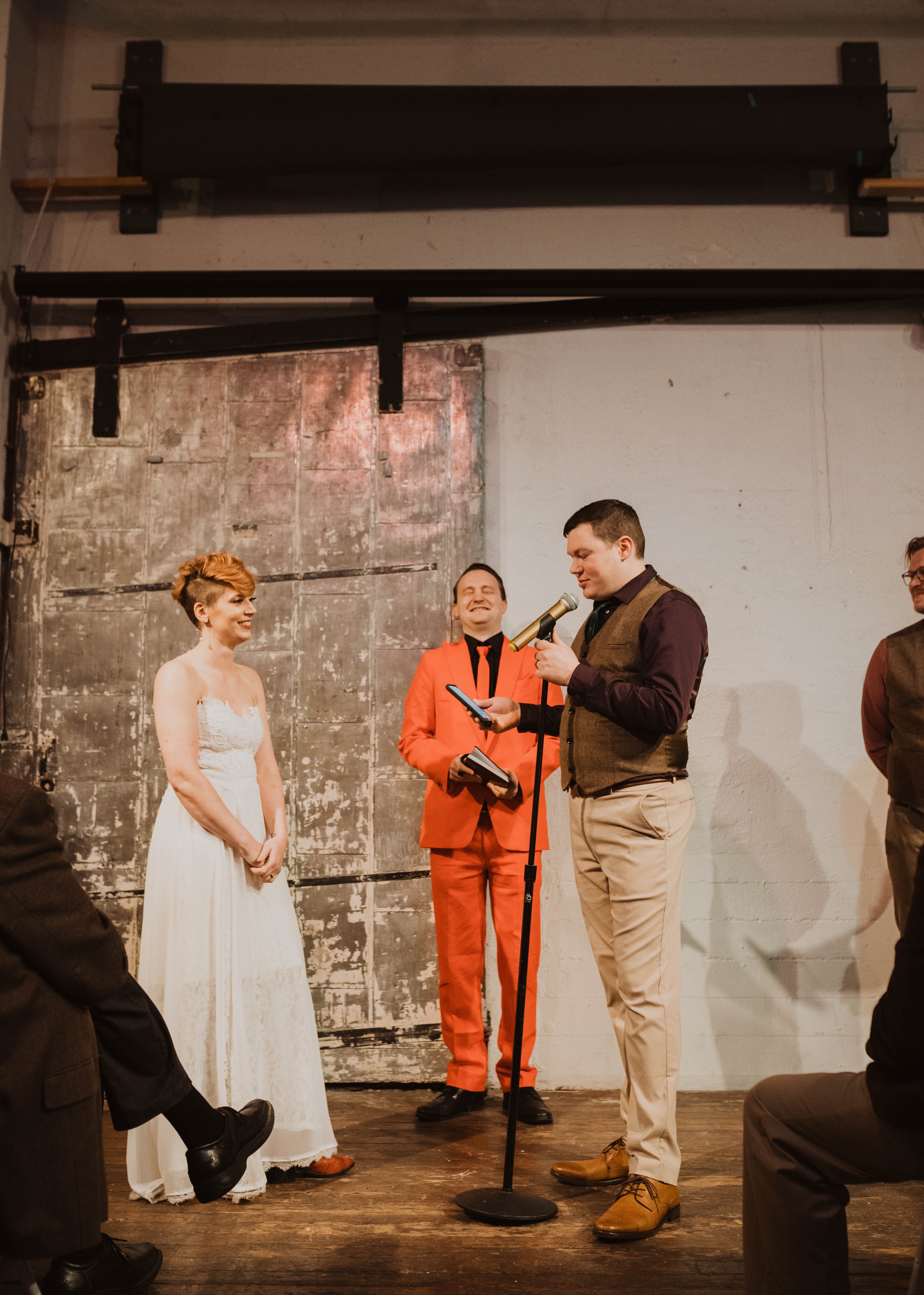 A wedding ceremony with a woman in a white dress and a man in a brown vest reading vows from a phone, standing in front of a rustic background with a person in a bright orange suit smiling.