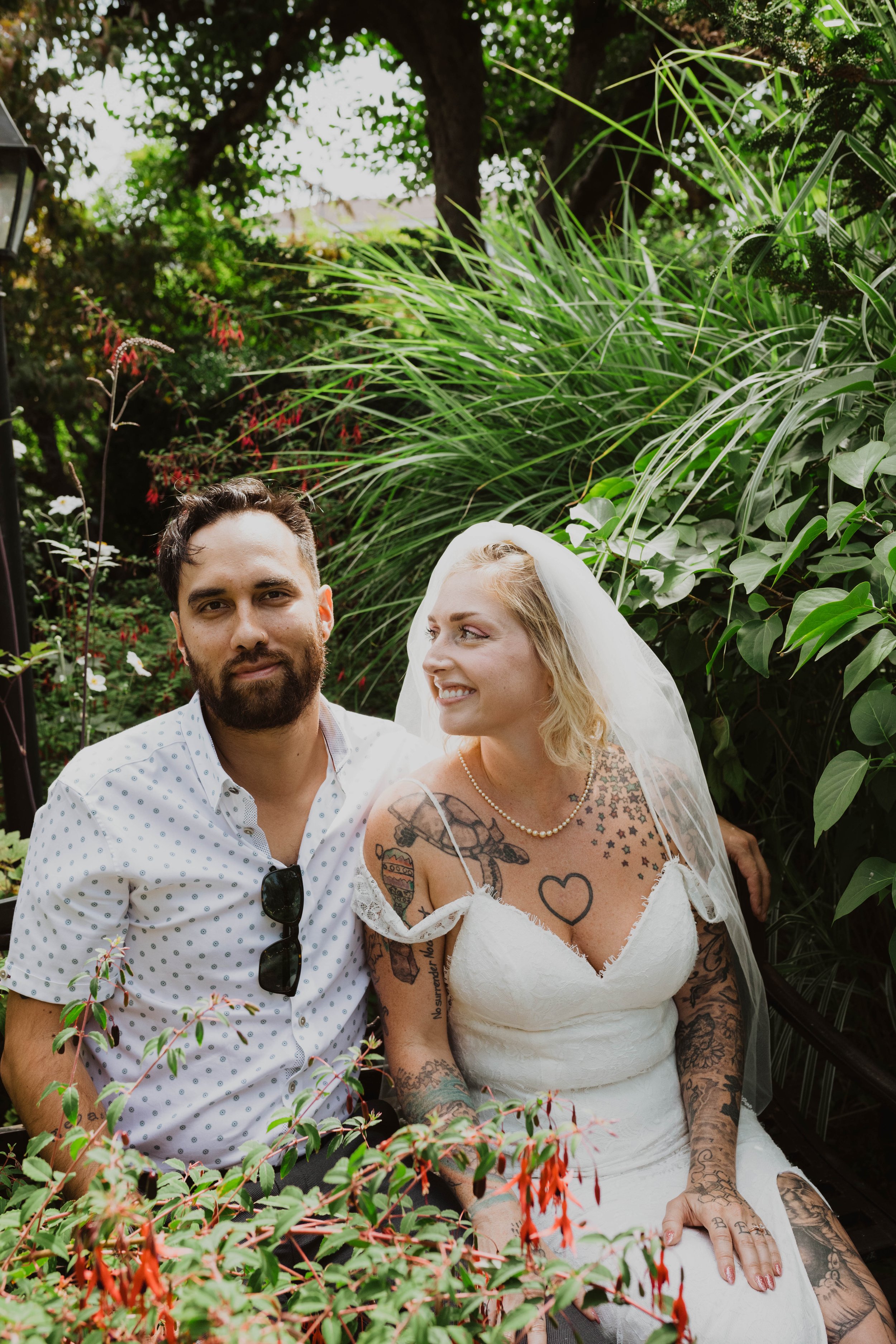 A newlywed couple sitting together outdoors surrounded by lush green plants. The man has dark hair, a beard, and is wearing a white polka-dot shirt with sunglasses hanging from his collar. The woman has blonde hair, tattoos on her arms and chest, a p