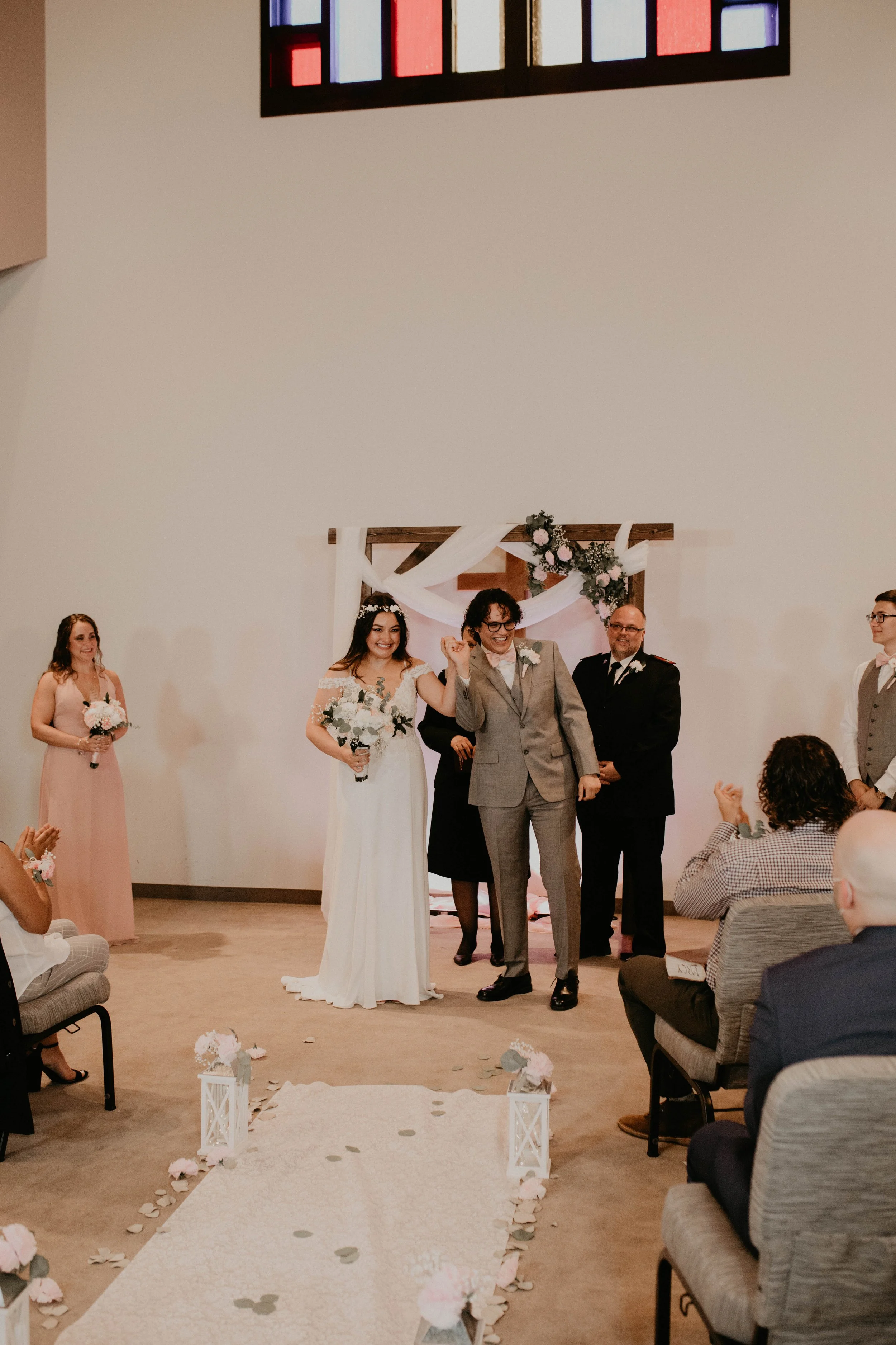 A wedding ceremony with a bride, groom, officiant, and guests inside a church or chapel. The bride is holding a bouquet, and the groom is holding her hand. The ceremony arch is decorated with flowers and white fabric. Guests are seated on chairs, wat