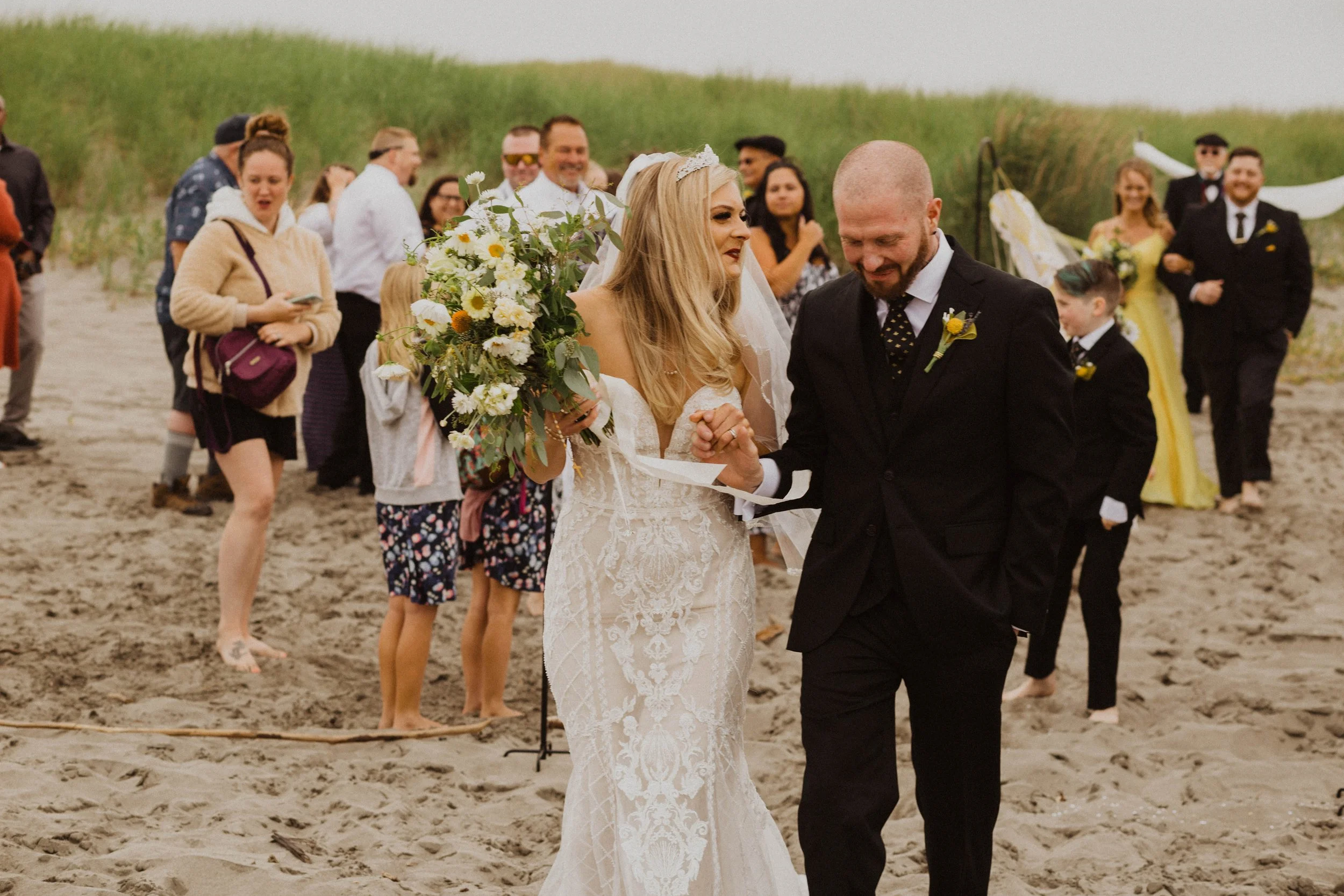 A wedding ceremony on the beach with the bride and groom holding hands, surrounded by guests. The bride wears a lace wedding dress, and the groom wears a black suit. Guests are dressed casually and formally, some taking photos. Long Beach, WA wedding