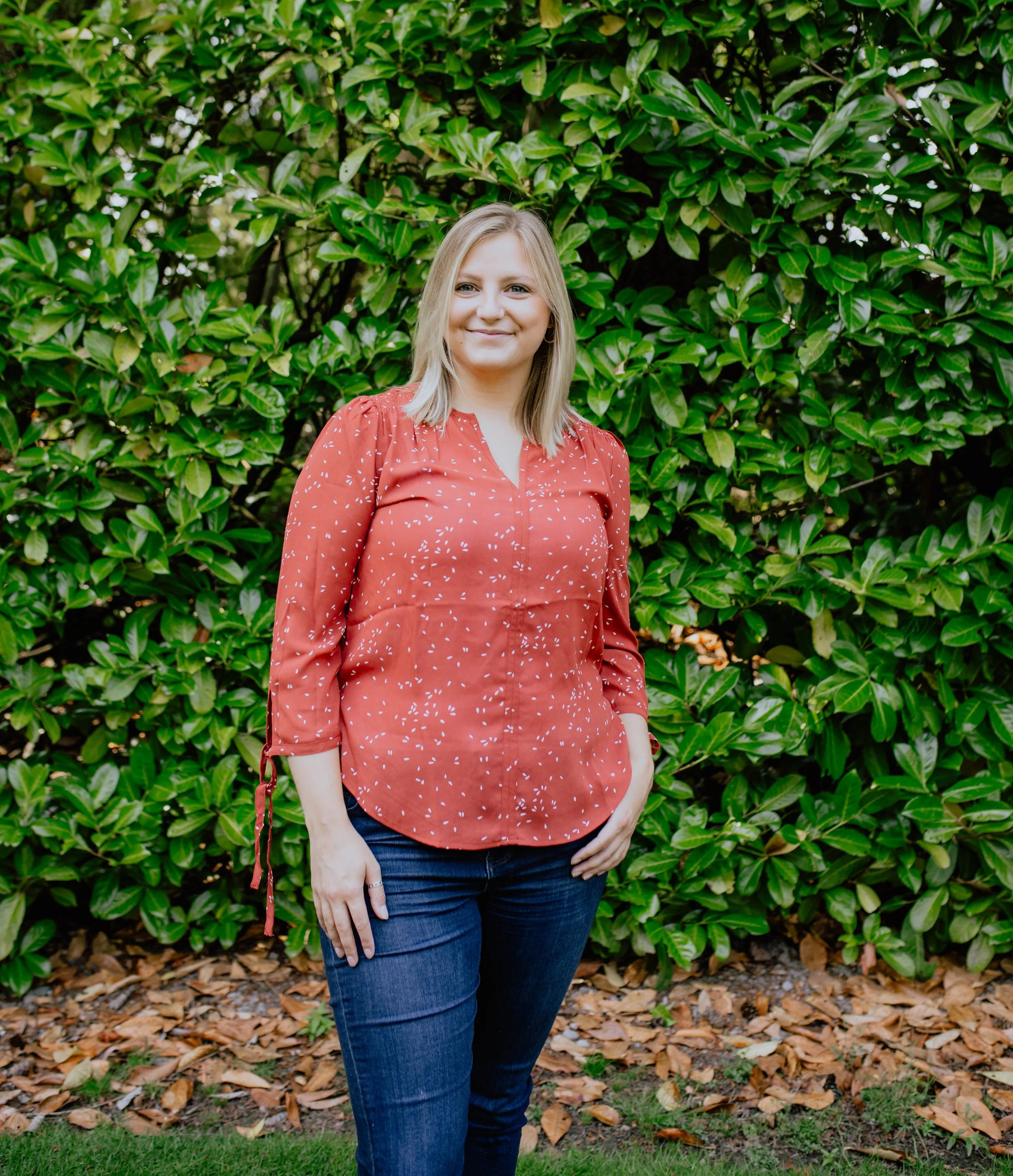 A woman standing outdoors in front of a lush green leafy bush, wearing a red patterned blouse and blue jeans, smiling at the camera. Seattle professional head shot photography
