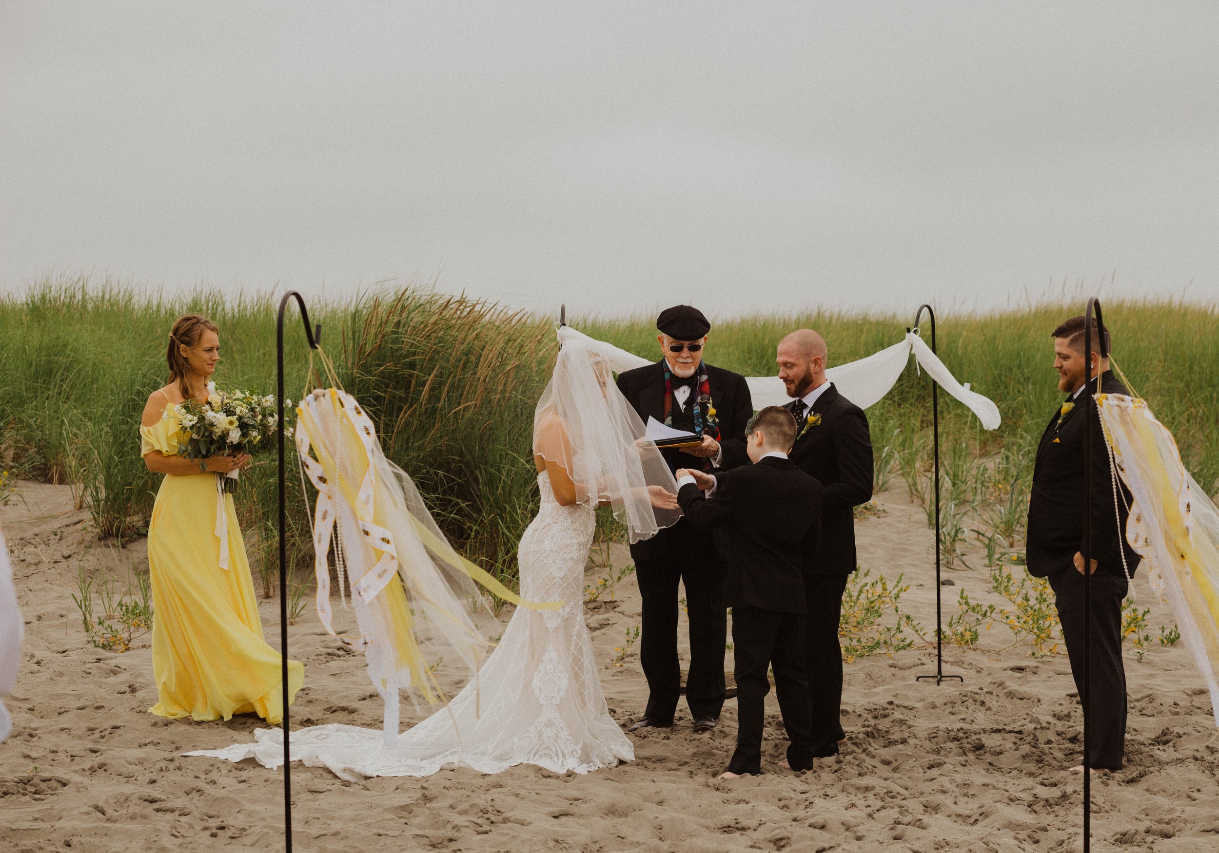 A beach wedding ceremony with a bride and groom, officiant, two boys, and two men, with some greenery and sandy ground. Long Beach, WA wedding photography.
