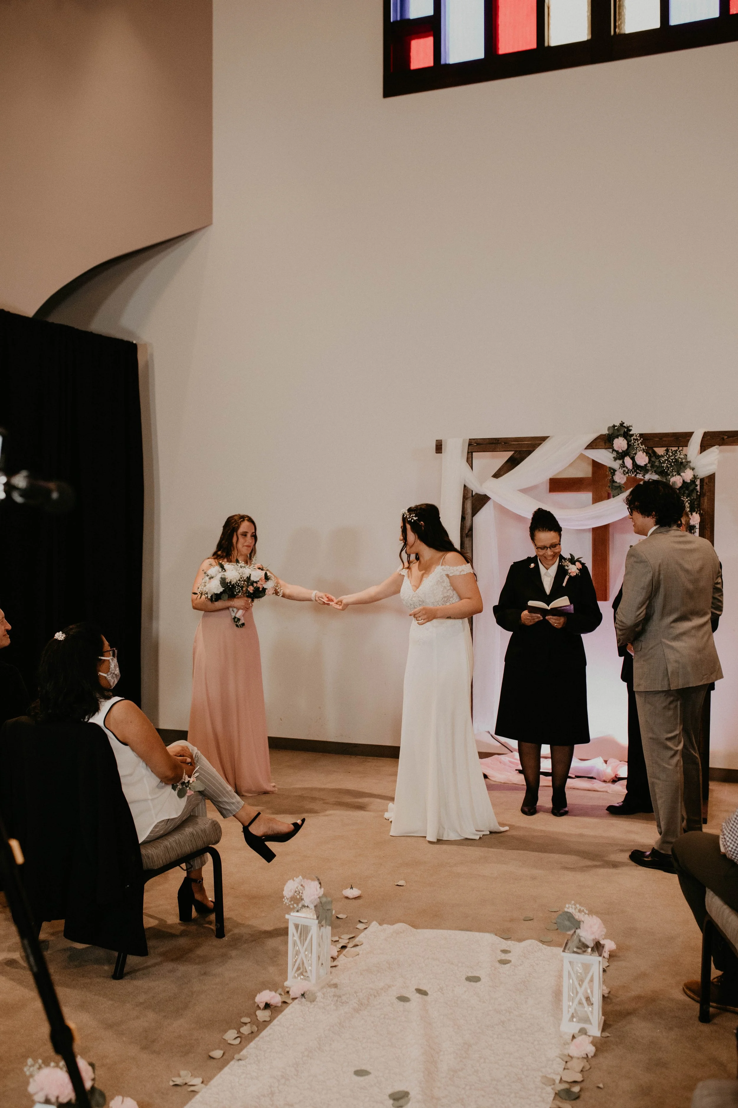 A wedding ceremony taking place inside a church, with two women holding hands, exchanging vows or rings, standing beneath a wooden arch decorated with flowers and white fabric, with a cross in the background, and a minister or officiant standing near