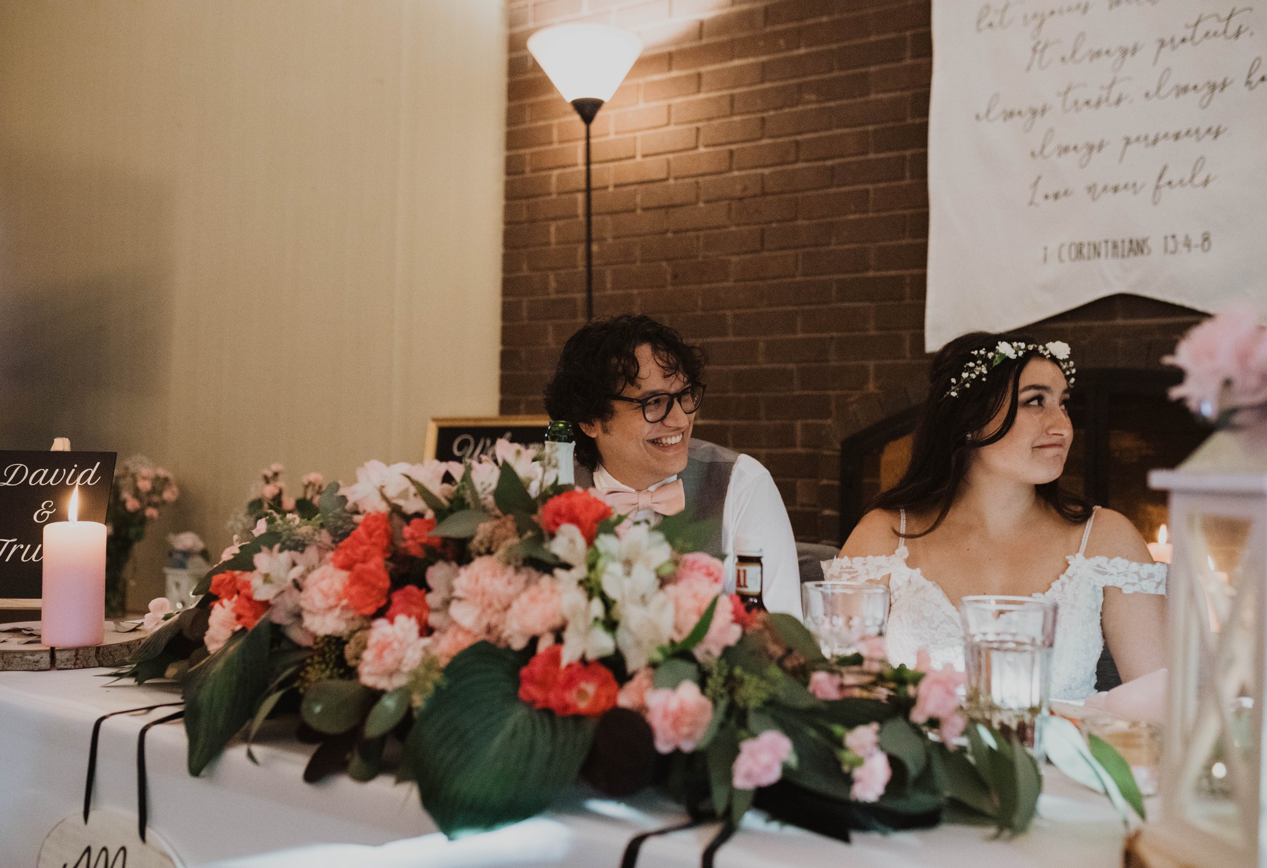 A groom and a bride sitting at a wedding reception table, decorated with pink and white flowers and greenery. The groom is smiling and wearing glasses, a gray vest, white shirt, and pink bow tie. The bride is wearing a white off-shoulder wedding dres