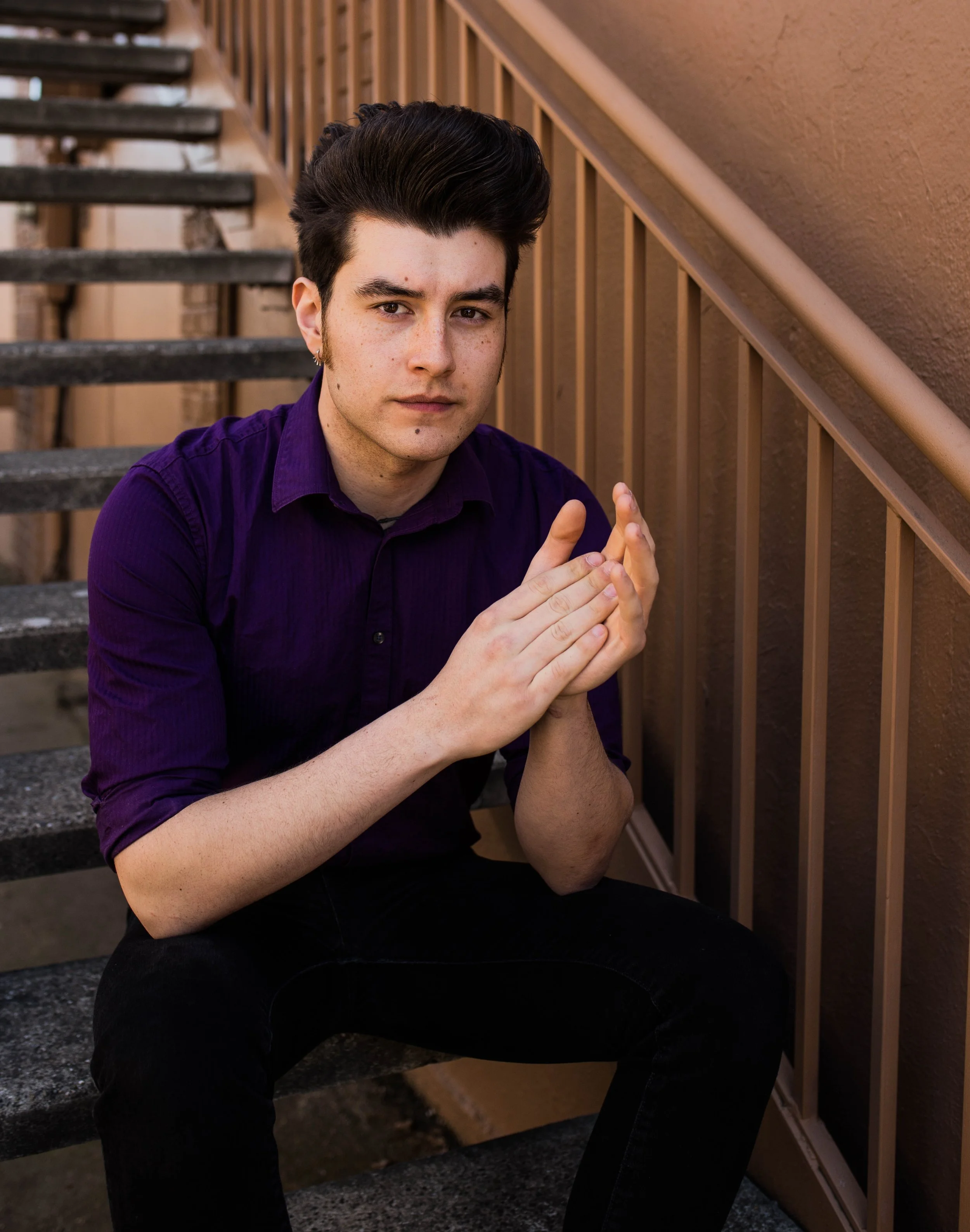 Young man sitting on outdoor staircase with hands together, wearing a purple shirt and black pants. Seattle professional head shot photography