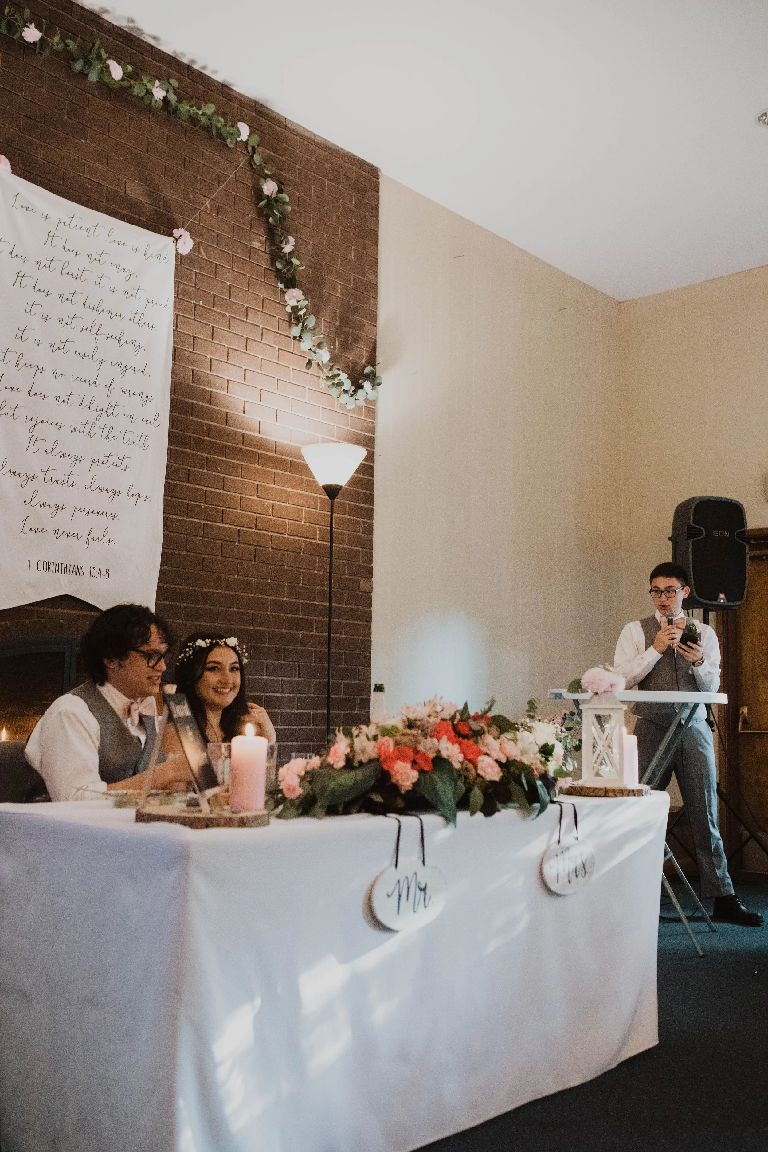 A wedding reception scene with two people sitting at a decorated table and a man standing at a microphone. The table is adorned with pink flowers and candles, and signs reading 'Mr.' and 'Mrs.' are hanging on the front. The backdrop features a brick 