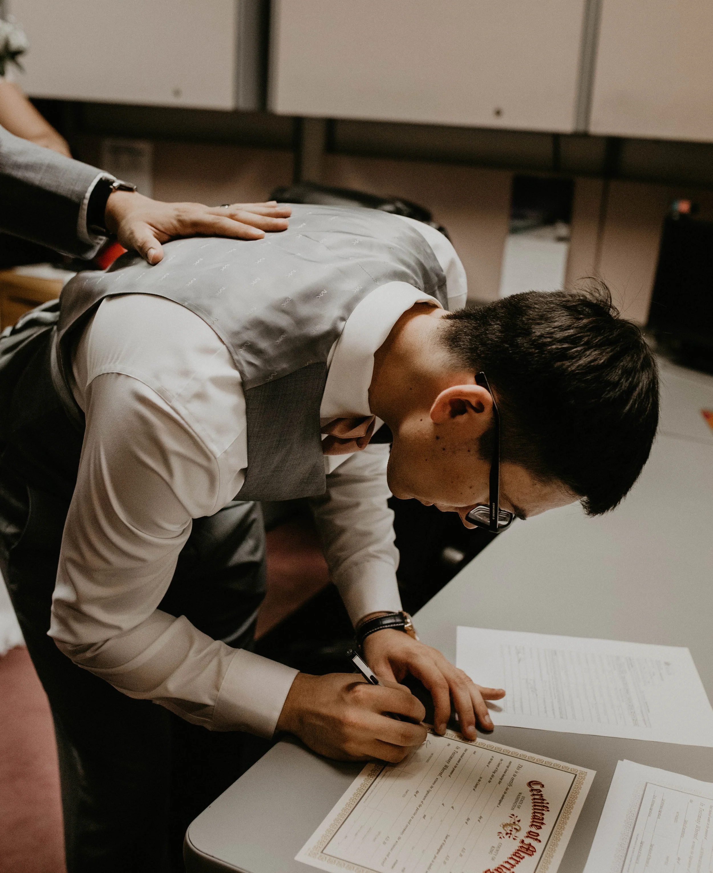 A man in a white shirt and gray vest signing a document on a desk with papers nearby, another person is placing a hand on his back. Seattle, WA wedding photography.