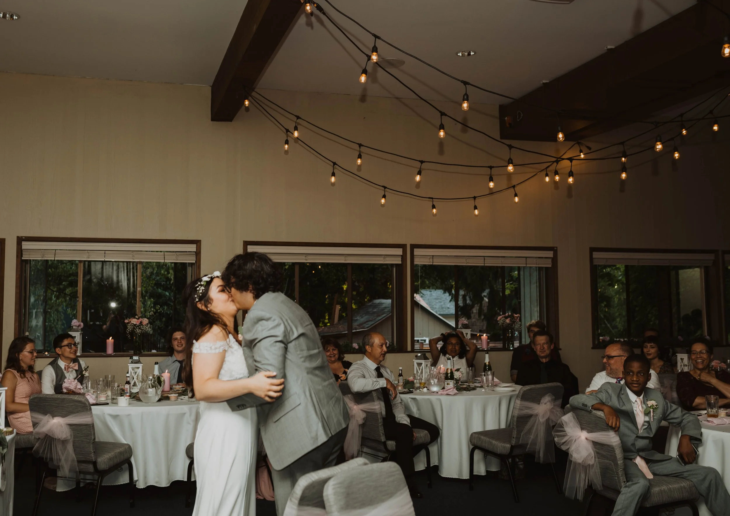 Bride and groom sharing a kiss at a wedding reception. Guests seated at round tables decorated with pink and white accents watch and smile. String lights hang from the ceiling, and windows with greenery outside are visible in the background.