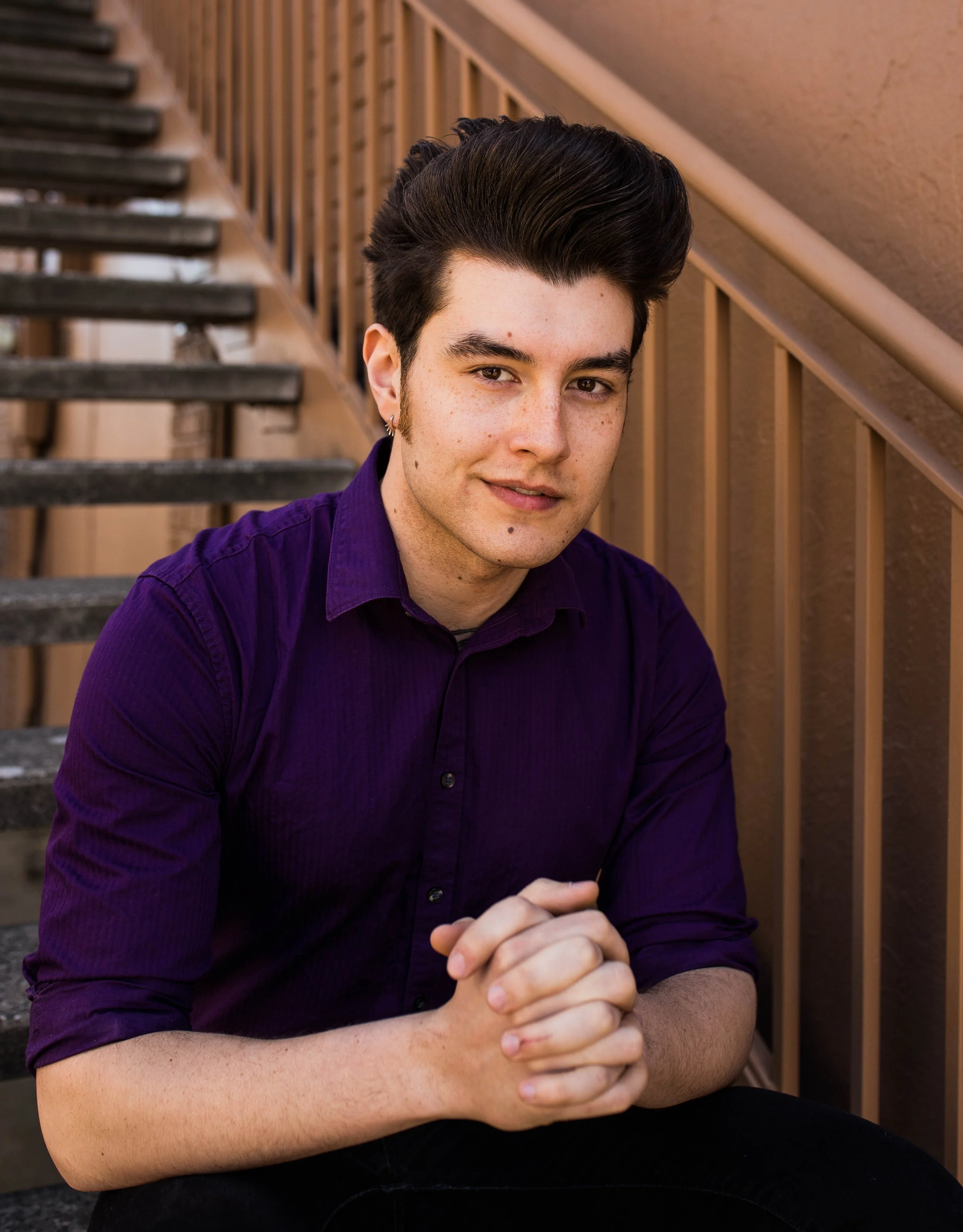 A young man with dark hair and earrings sitting on outdoor stairs, wearing a purple shirt, with his hands clasped. Seattle professional head shot photography