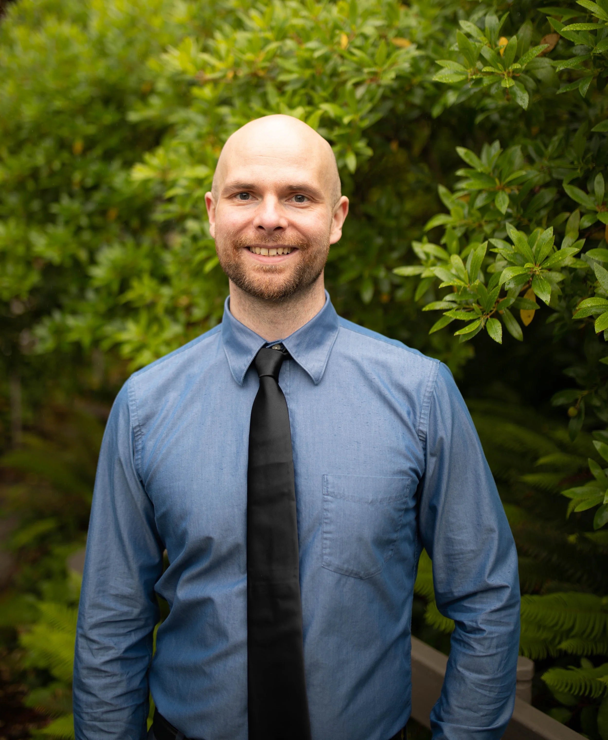 A smiling man with a beard and a bald head wearing a blue dress shirt and a black tie, standing outdoors in front of green plants. Seattle professional head shot photography