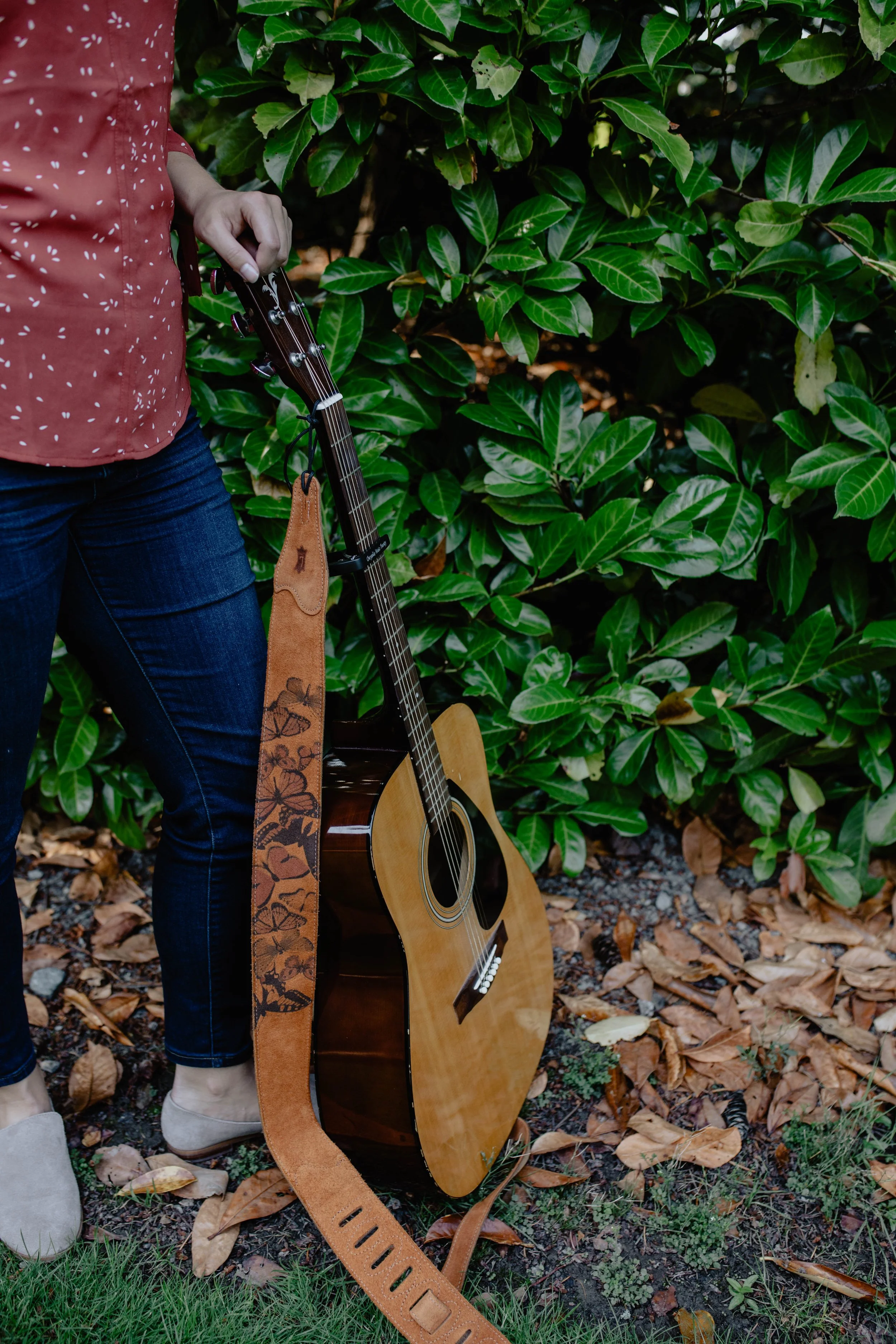 Person holding an acoustic guitar with a leather strap, standing next to bushes and fallen leaves. Seattle professional head shot photography