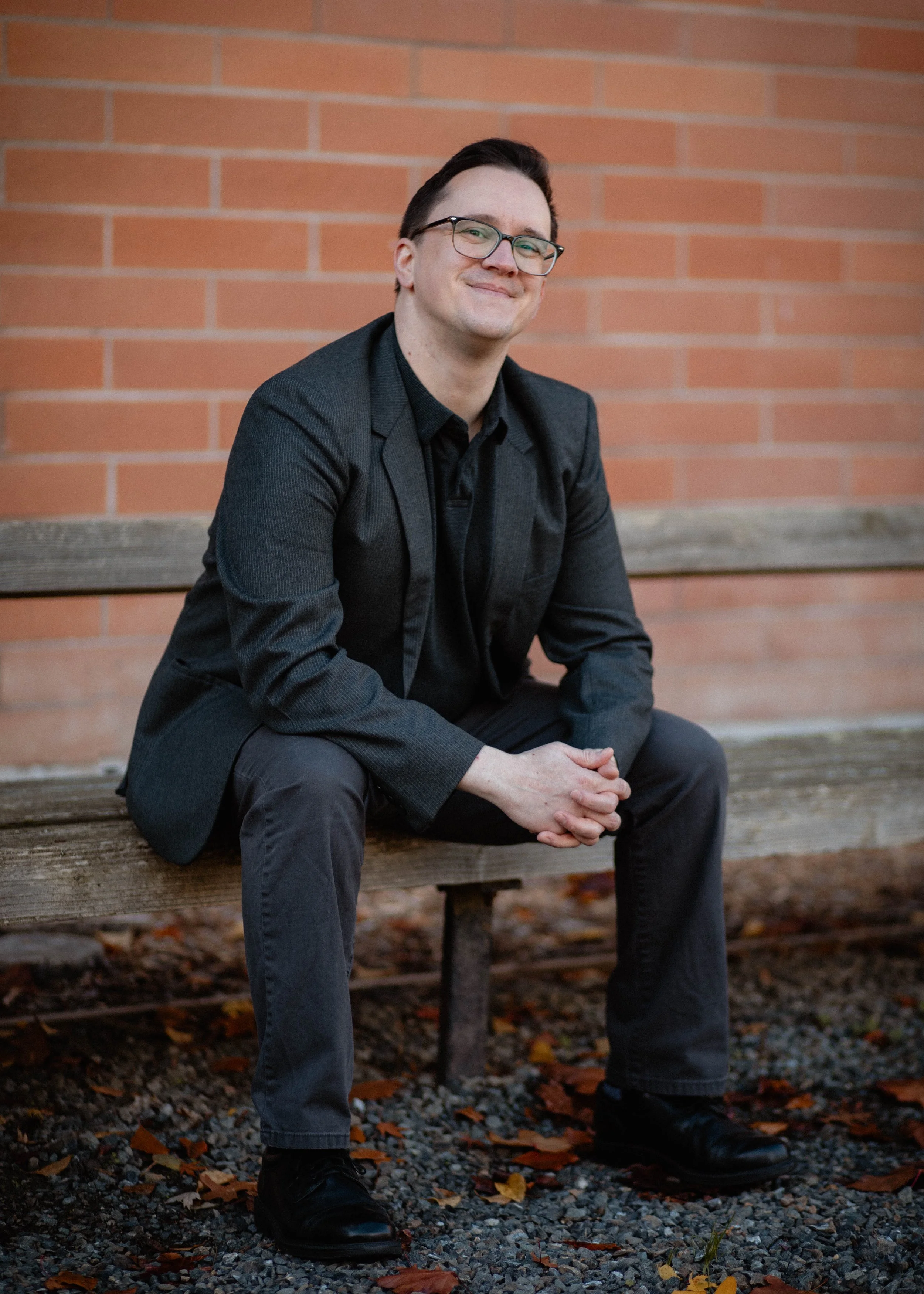 A man wearing glasses, a dark blazer, and dark pants sitting on a wooden bench outdoors in front of a brick wall, with autumn leaves on the ground. Seattle professional head shot photography