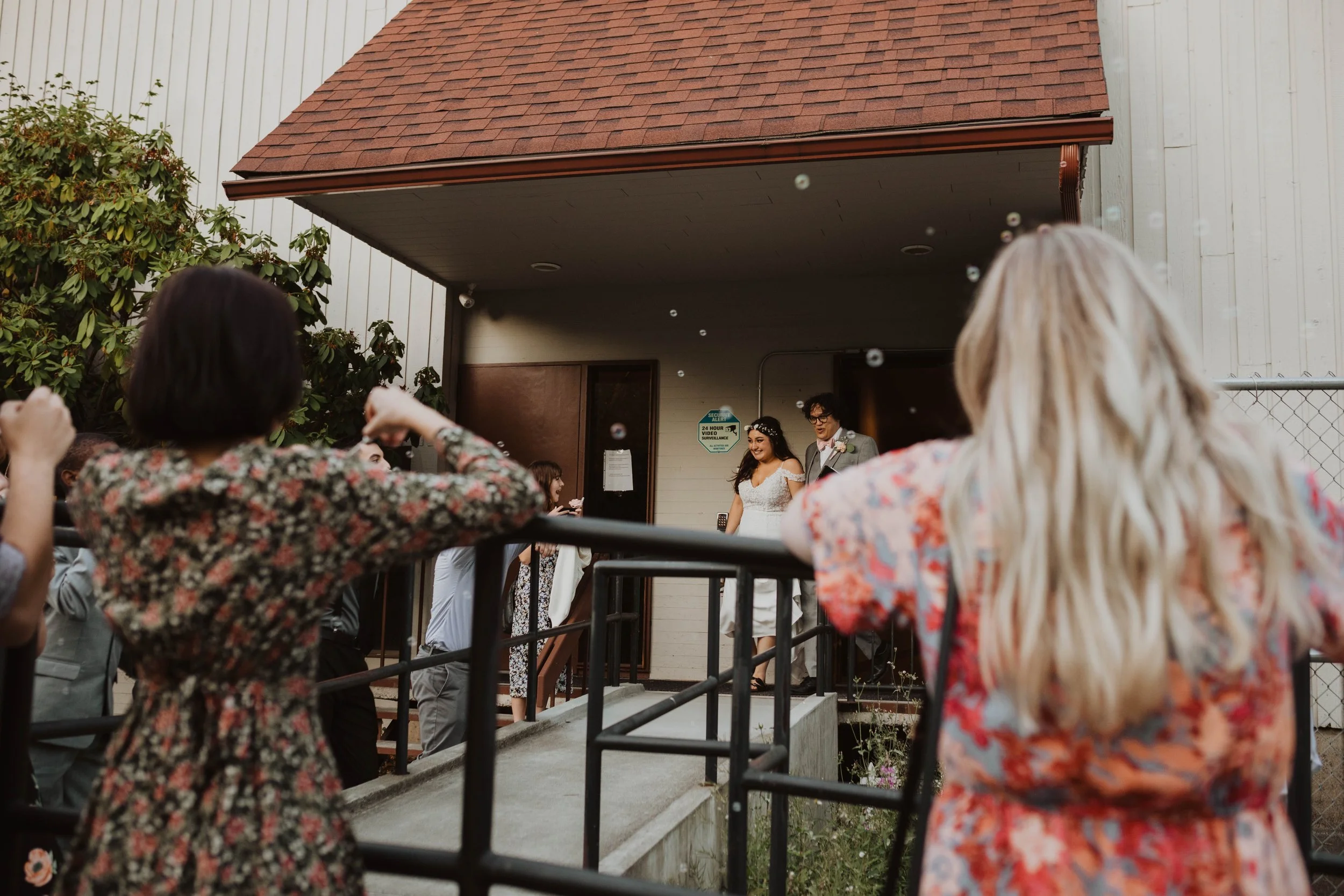 A wedding ceremony with a bride and groom standing in front of guests, outside a building with a red shingled roof, as guests throw bubbles in the air. Seattle, WA wedding photography.