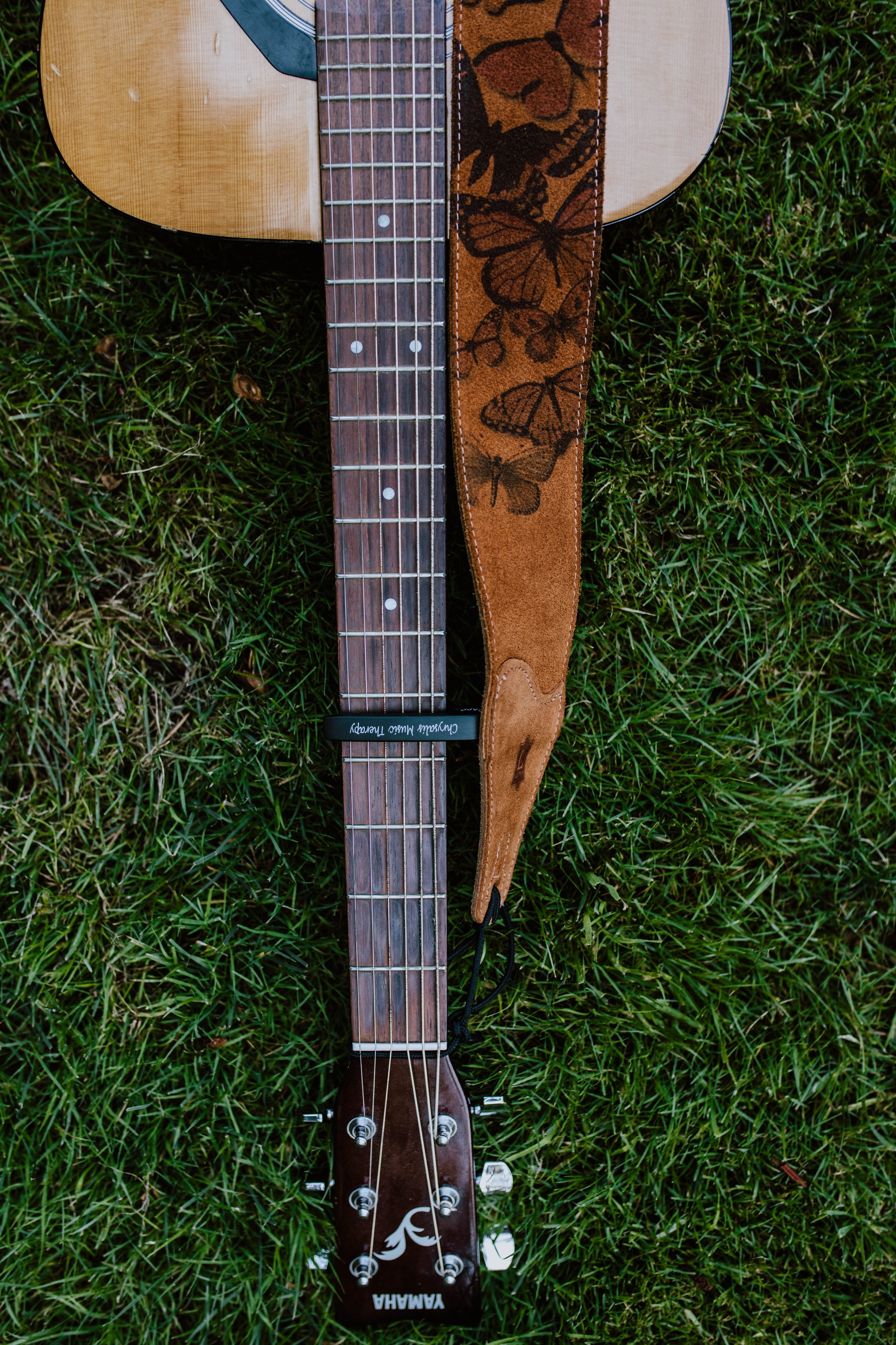 An acoustic guitar with a brown strap featuring butterfly and tiger designs lying on green grass. Seattle professional head shot photography