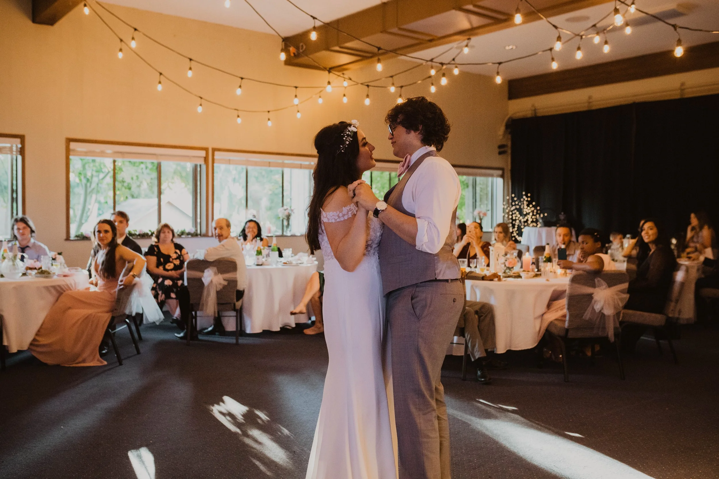 A bride and groom sharing their first dance at a wedding reception, inside a decorated hall with guests seated at tables in the background. Seattle, WA wedding photography.