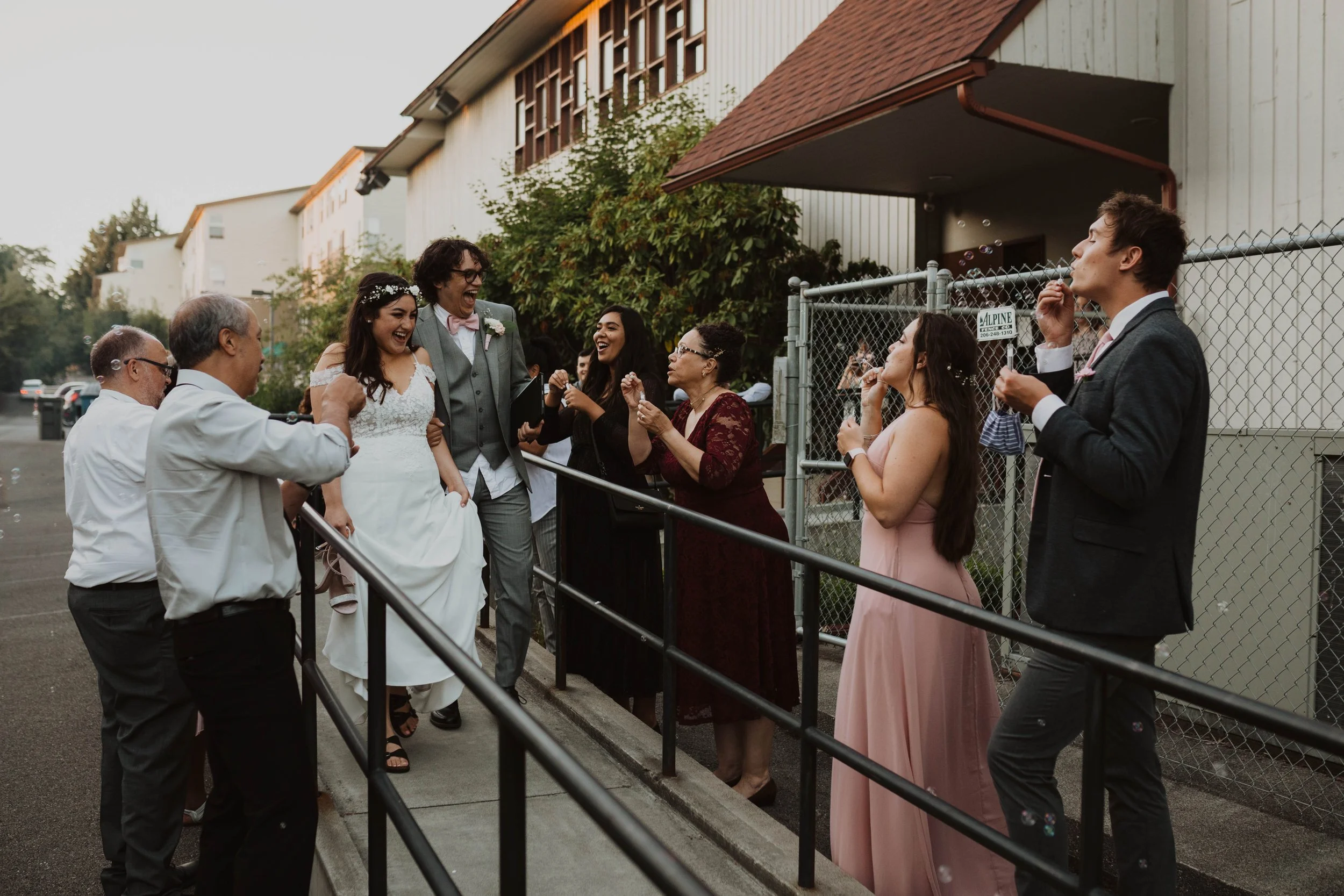 People celebrating outside a building, including a bride in a white wedding dress, a groom in a gray suit, and friends and family enjoying the moment. Seattle, WA wedding photography.