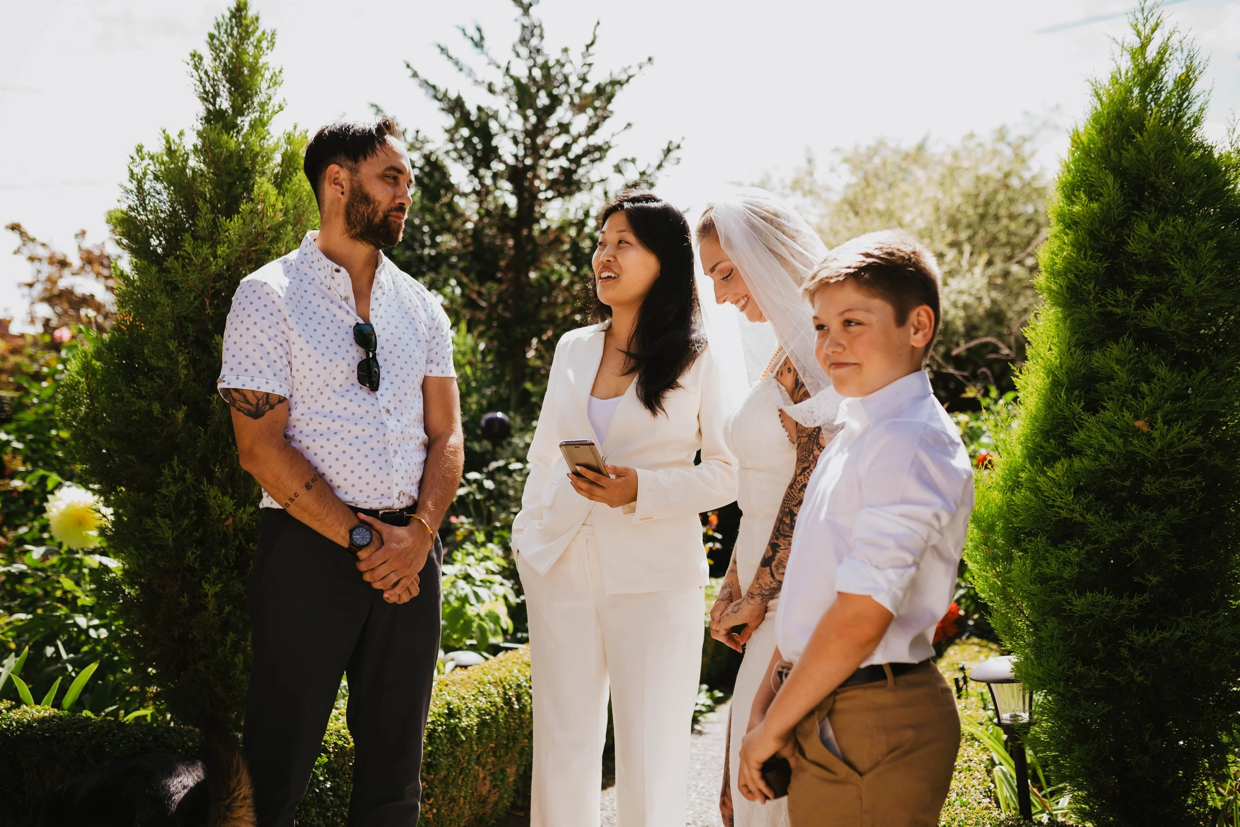 A group of five people gathered outdoors in a garden, including a woman in a wedding dress with a veil, and others dressed in formal attire, smiling and engaging in conversation on a sunny day. Seattle, WA wedding photography.