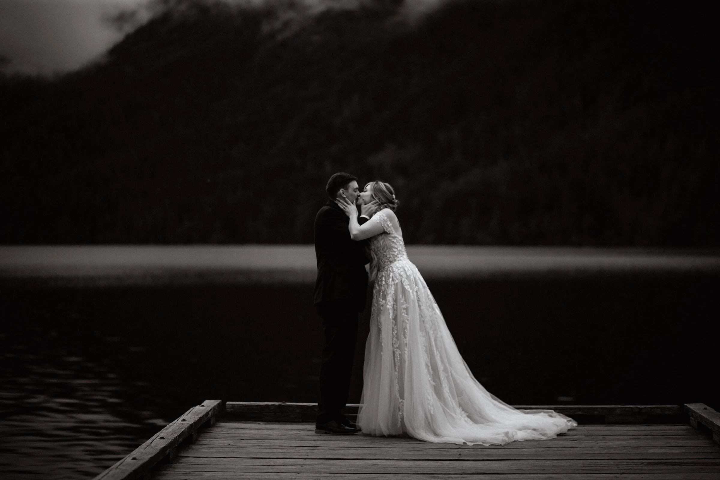 Bride and groom share a passionate kiss on the dock of Lake Crescent in Port Angeles, WA after exchanging their vows. The image is in black and white, cinematic landscape