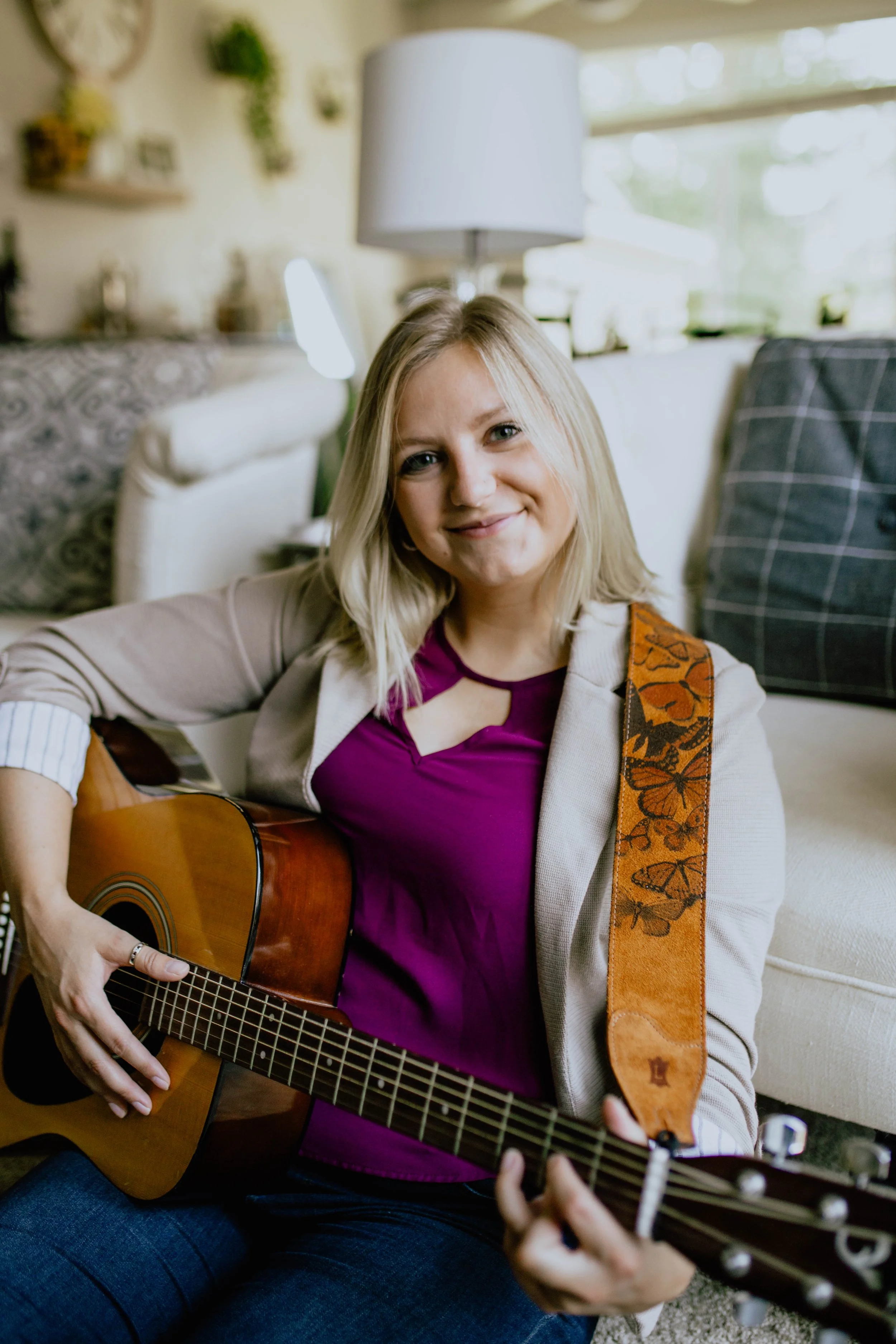 A woman with blonde hair smiling while sitting on the floor playing an acoustic guitar in a cozy living room. Seattle professional head shot photography