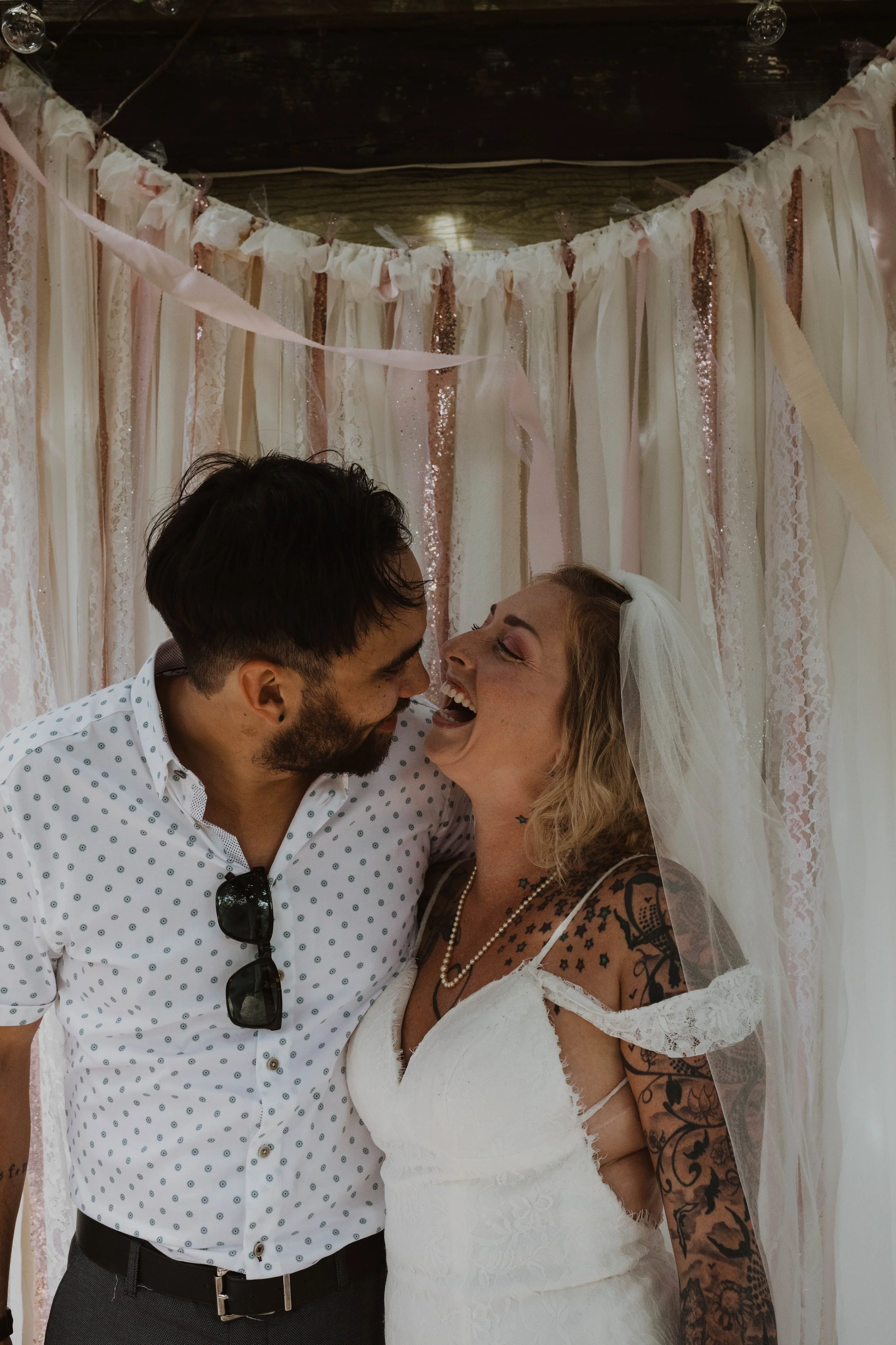 A couple celebrating a wedding, with the woman wearing a wedding dress and veil, and the man in a white shirt with sunglasses hanging from the collar, standing in front of a backdrop decorated with pink, white, and glittery ribbons.