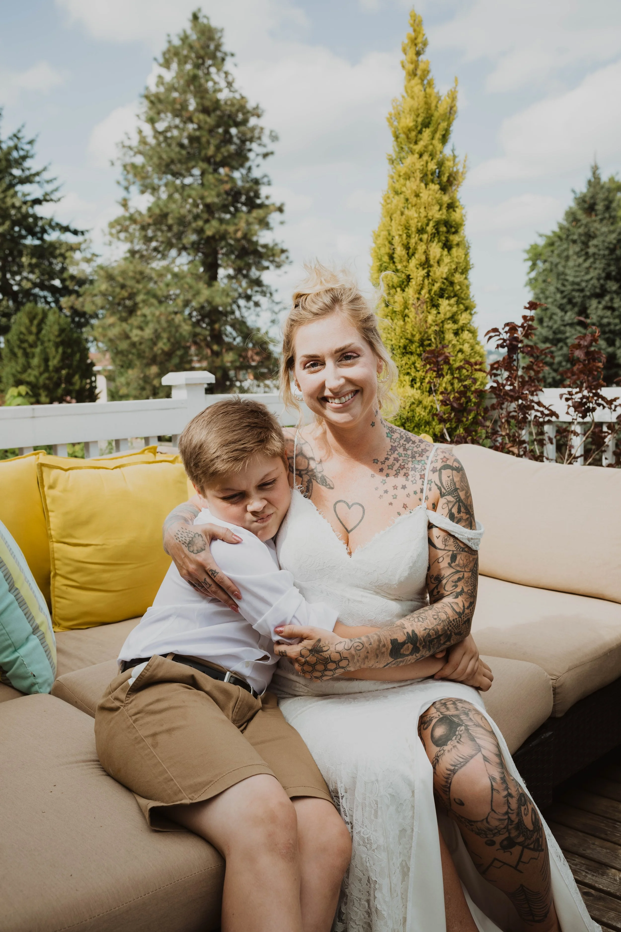 A woman with blonde hair and tattoos on her arms and legs is smiling while holding a young boy with brown hair, who appears to be making a playful face. They are sitting on an outdoor beige sofa with colorful cushions, against a backdrop of trees and