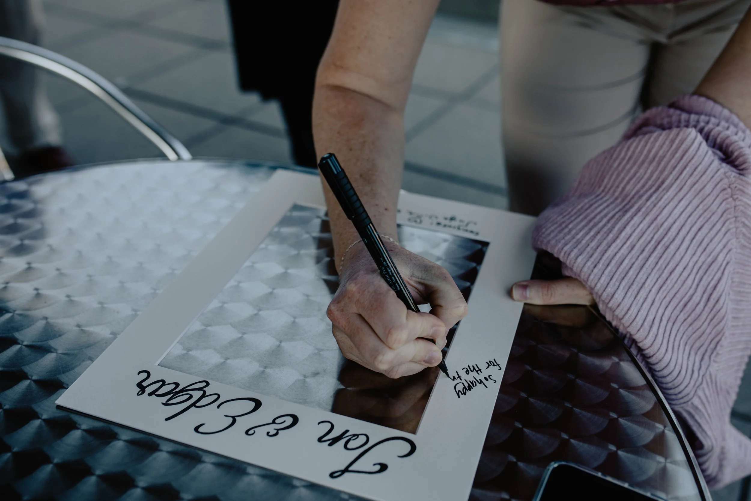 A person signing a photo at an outdoor table with a black marker, wearing a pink sweater. Seattle Municipal Courthouse wedding photography.