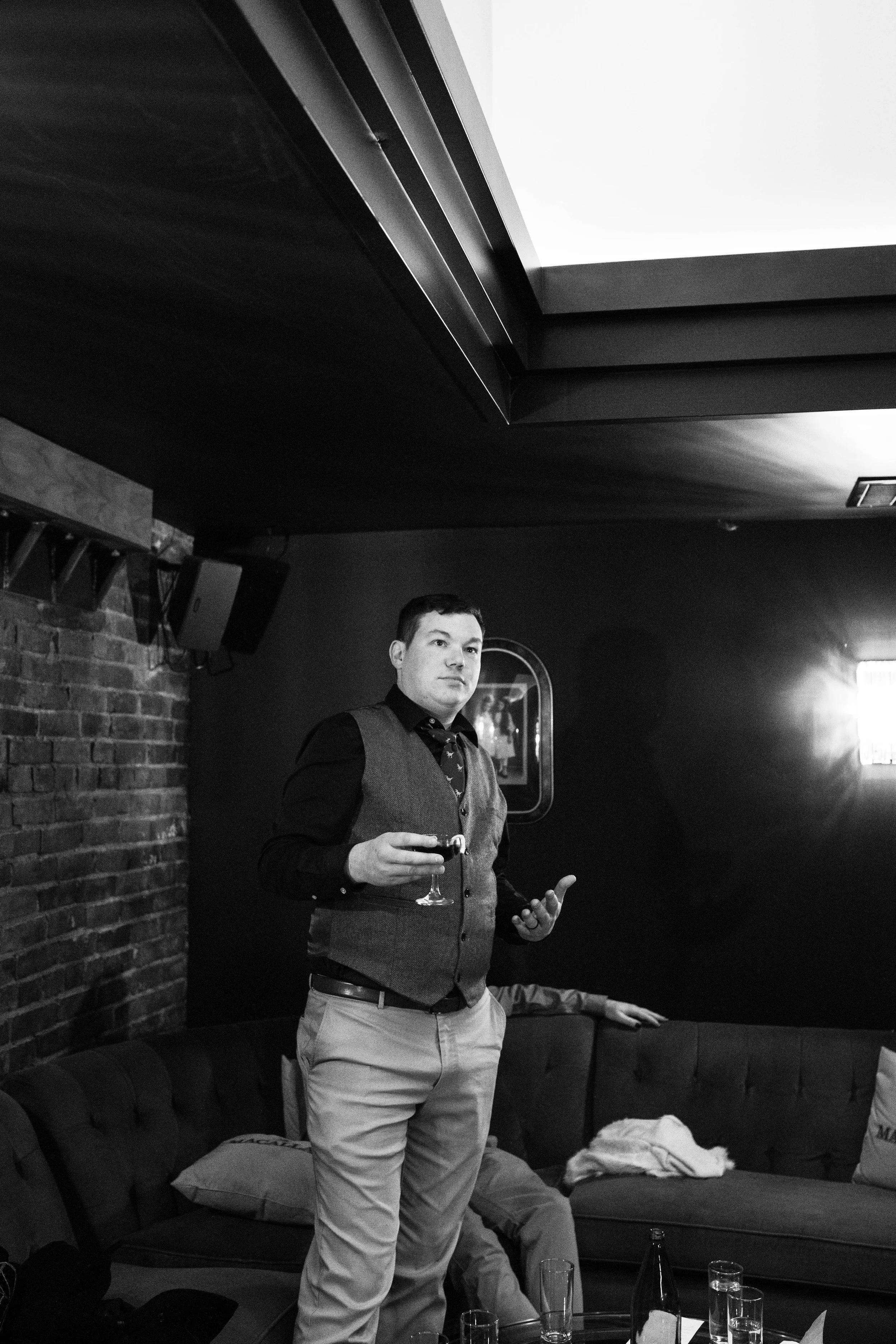 A man holding a glass of wine standing in a dimly lit room with a brick wall, dark ceiling, and a couch, with sunlight coming through a window. Pioneer Square, Seattle, WA wedding photography.