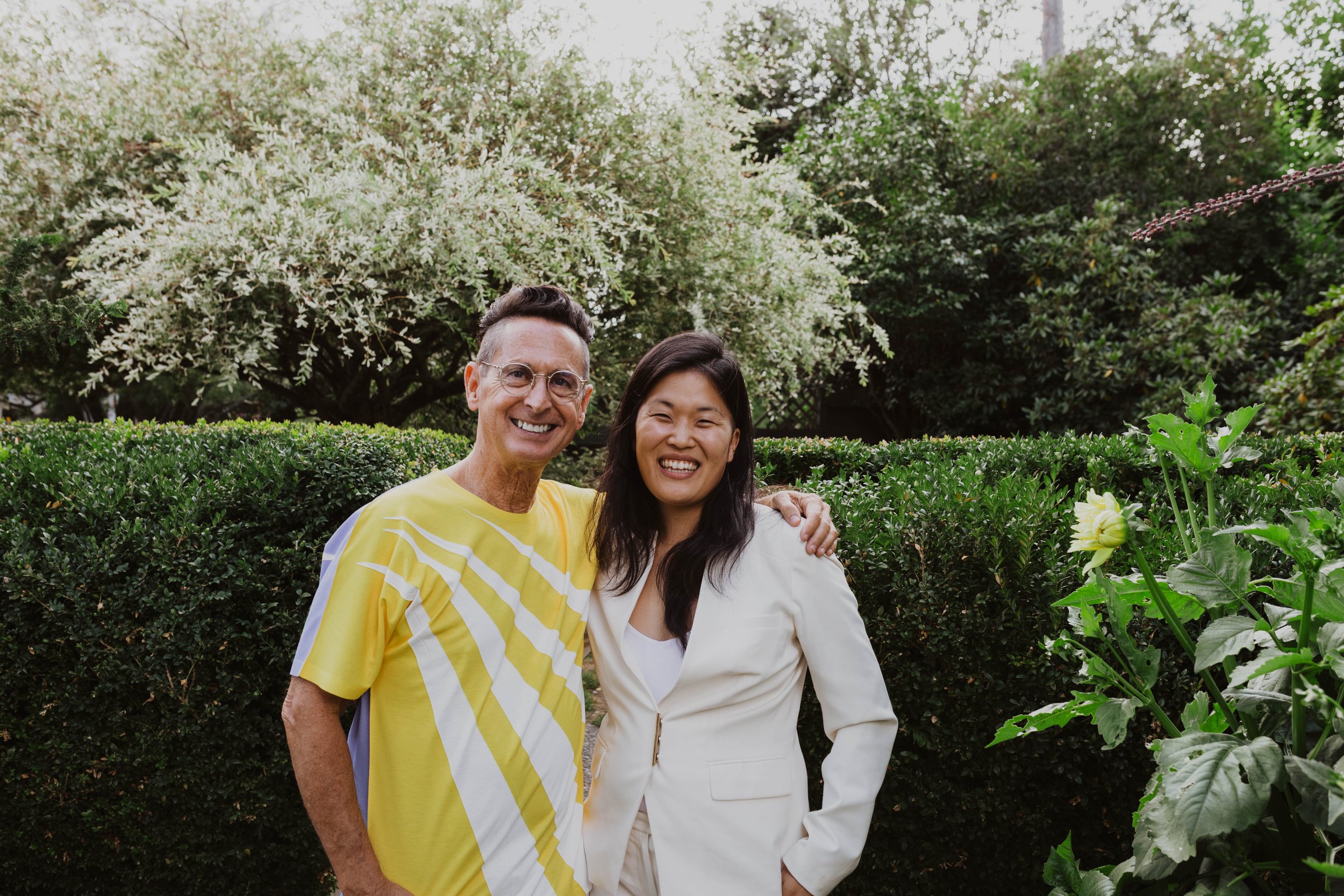 Two smiling people, one man and one woman, standing outdoors in front of green bushes and trees, with the man wearing a yellow shirt and the woman wearing a white jacket. Seattle, WA wedding photography.