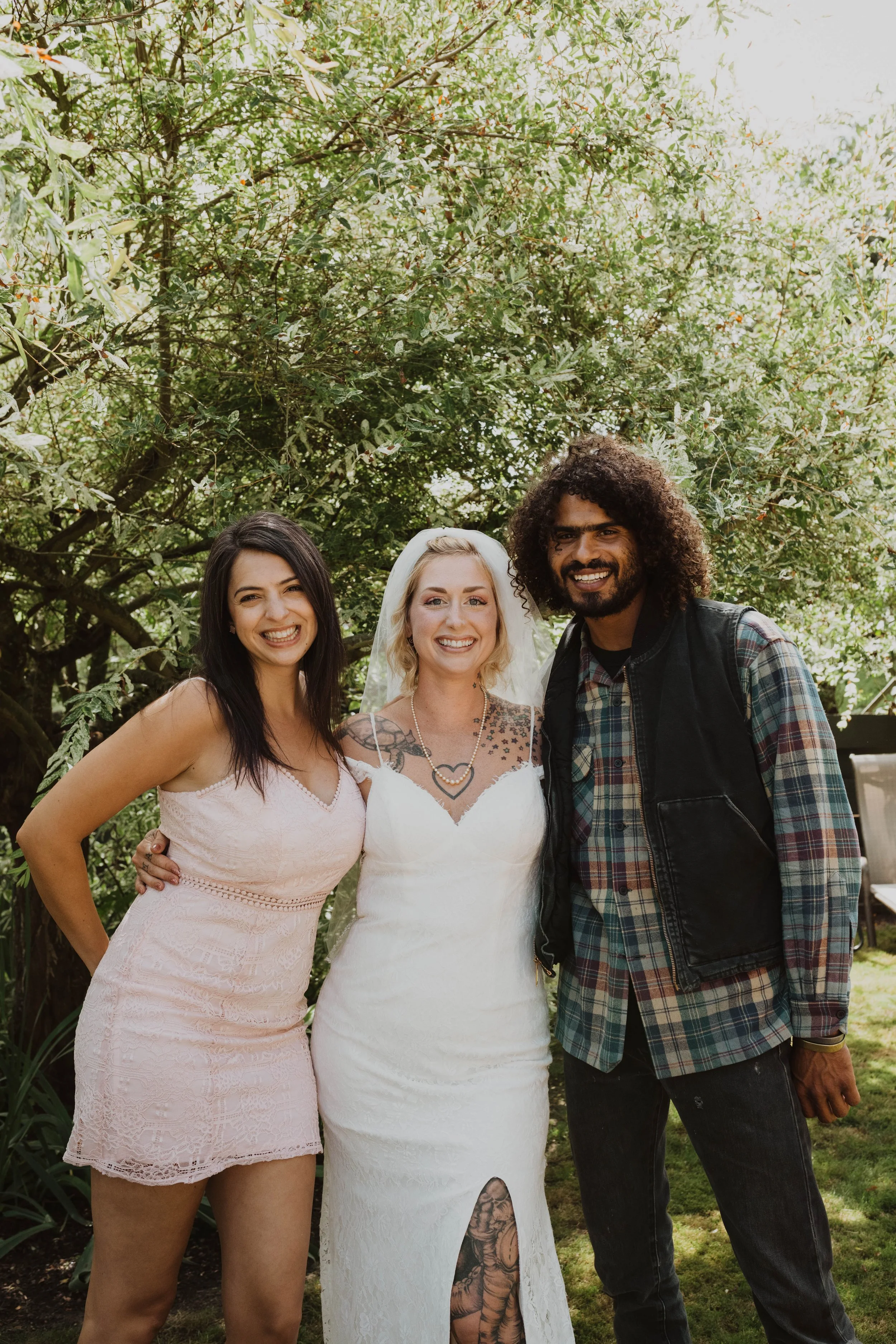 Three smiling people standing outdoors in front of green trees, two women and one man, dressed casually, celebrating a wedding. Seattle, WA wedding photography.