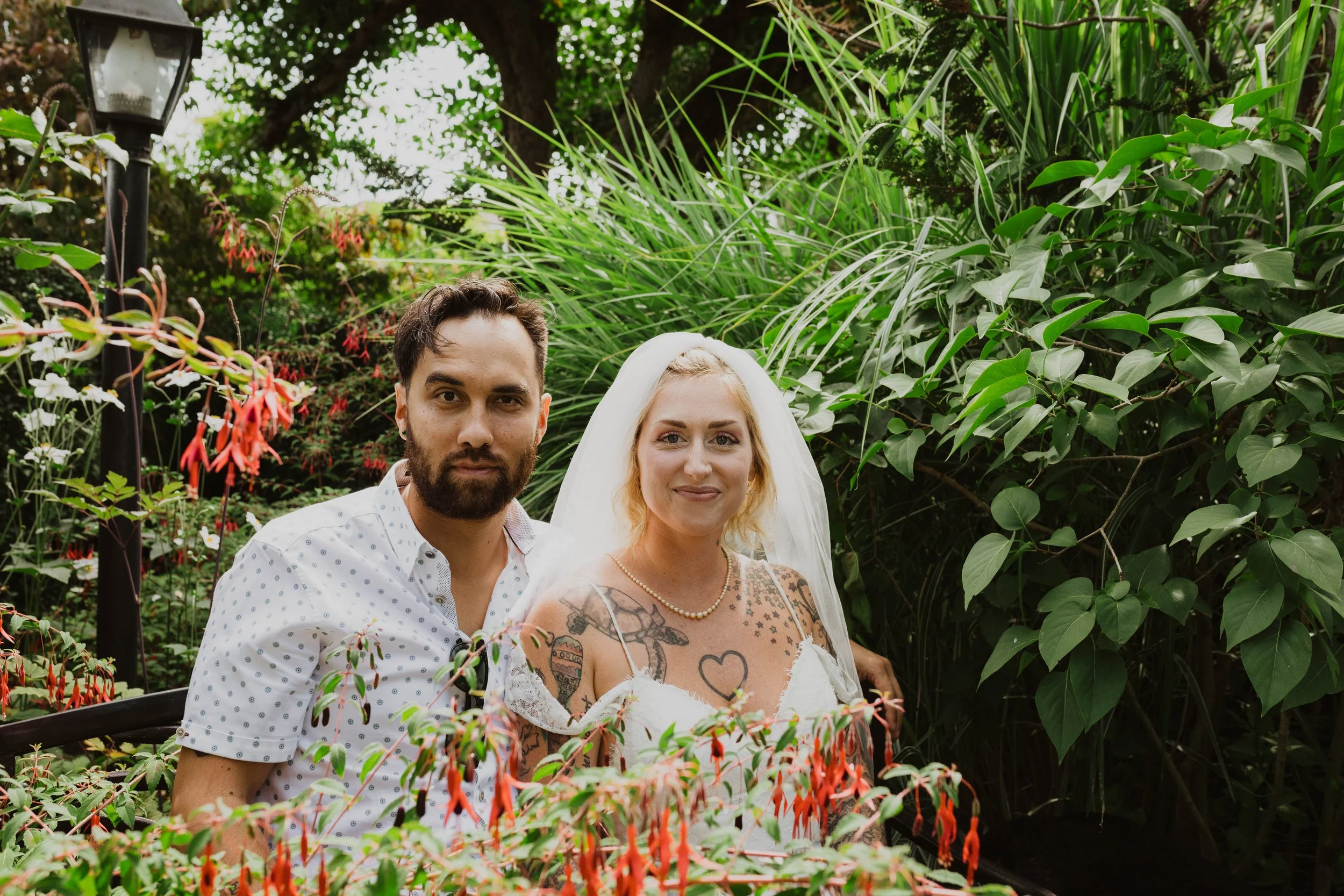 A couple, a man with dark hair and a beard, and a woman with blonde hair and tattoos, dressed in wedding attire, standing together in lush garden foliage with a lamppost visible in the background. Seattle, WA wedding photography.