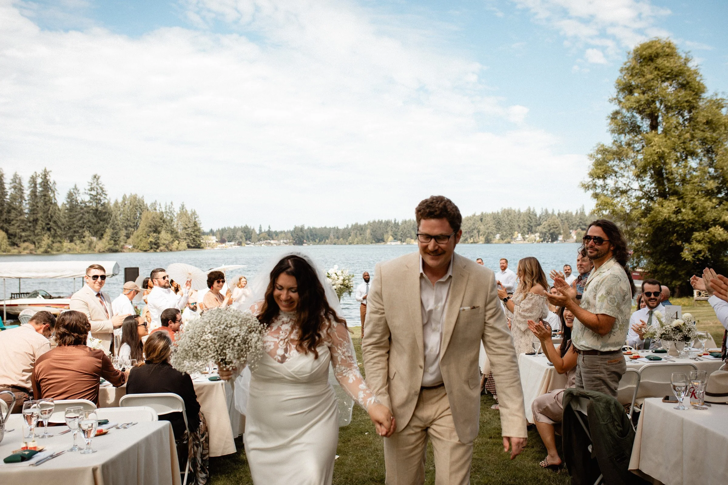 Bride and groom are holding hands as they walk back down the aisle as husband and wife. Their guests surrond them with love and the view of the lakeside in Seattle, WA