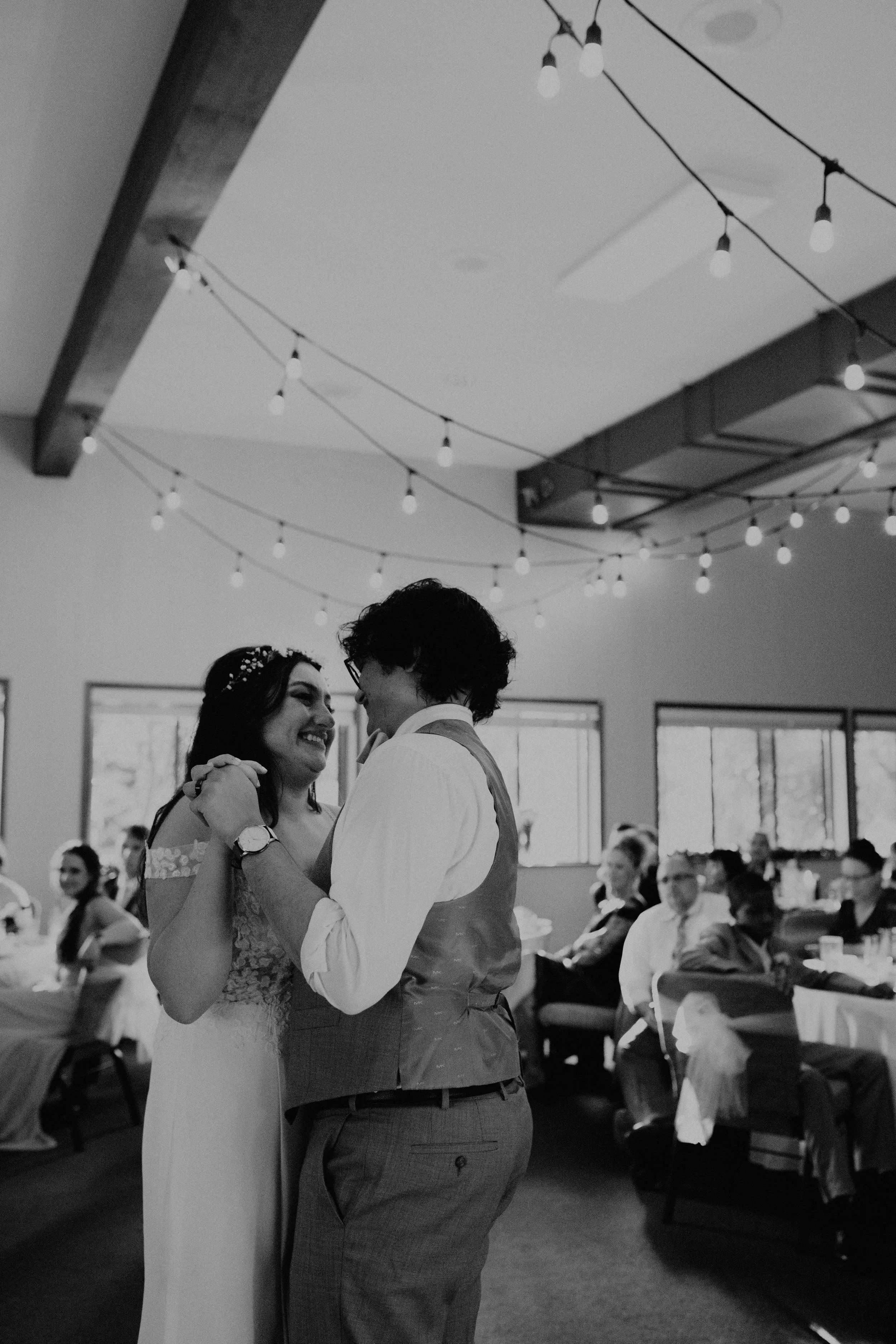 Black and white photo of a newlywed couple dancing at their wedding reception, surrounded by seated guests and decorated with string lights. Seattle, WA wedding photography.