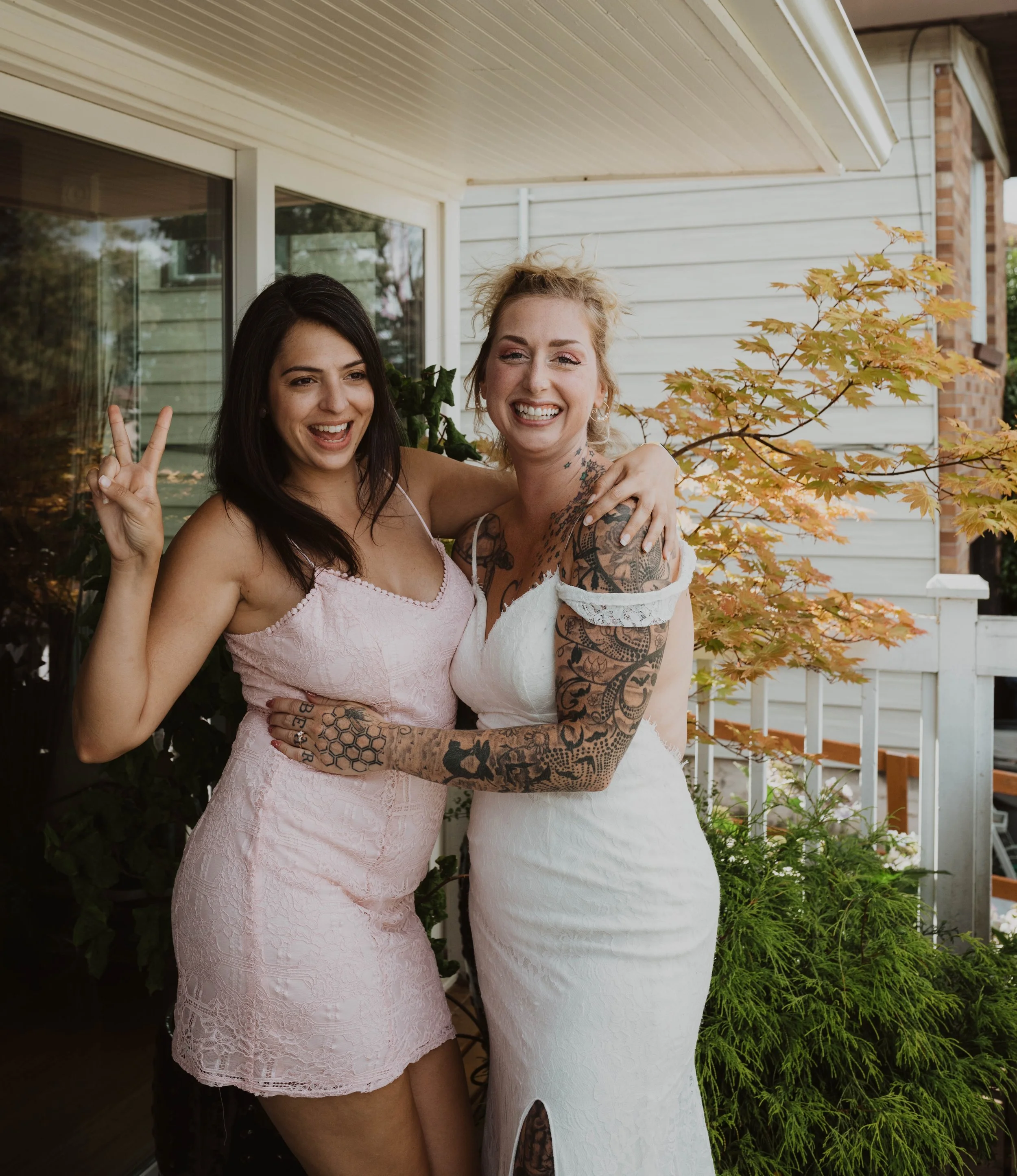 Two women in dresses smiling and posing together on a porch with autumn leaves and plants background. Seattle, WA wedding photography.