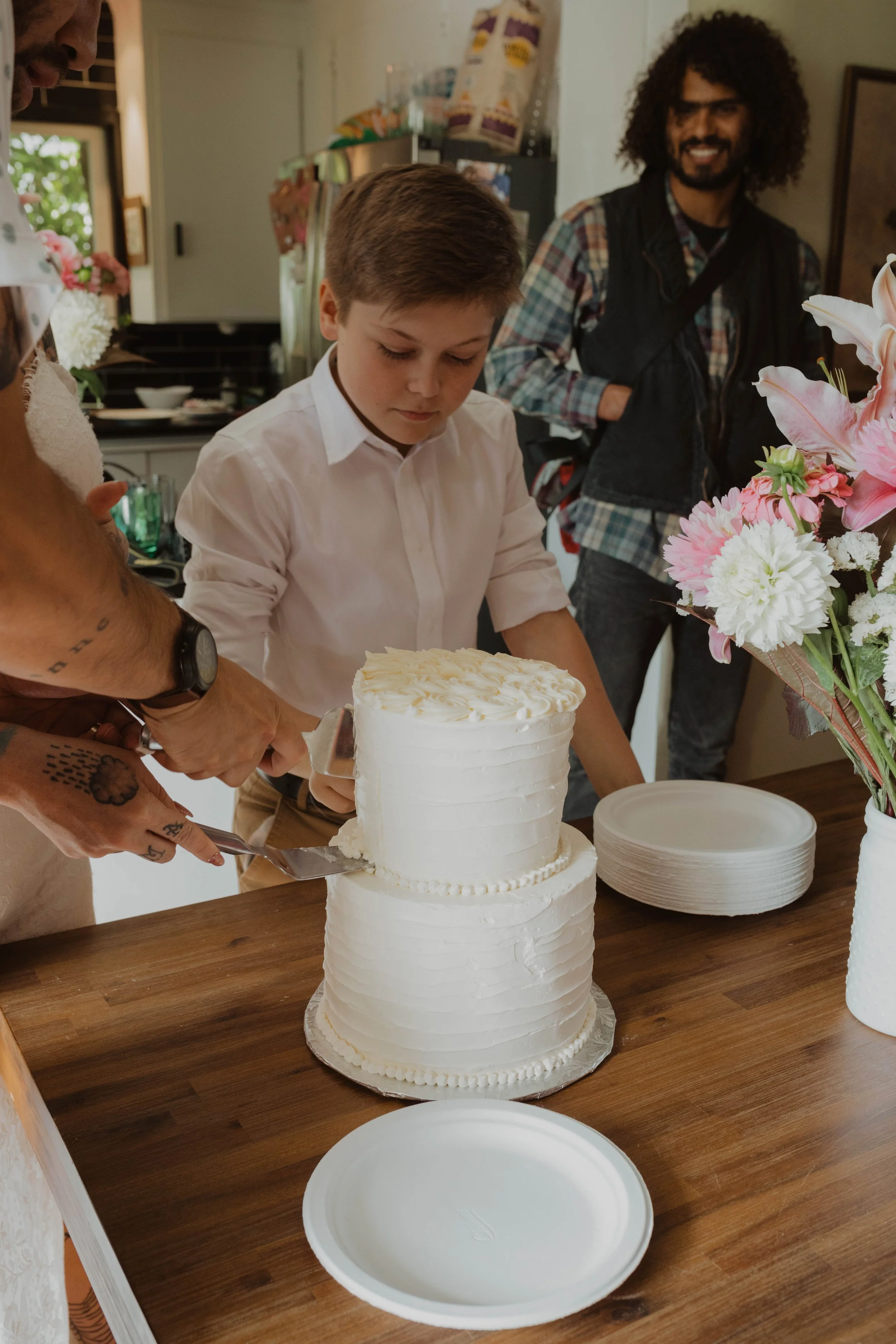 A boy is cutting a white wedding cake with people around him in a kitchen or dining room. Seattle, WA wedding photography.