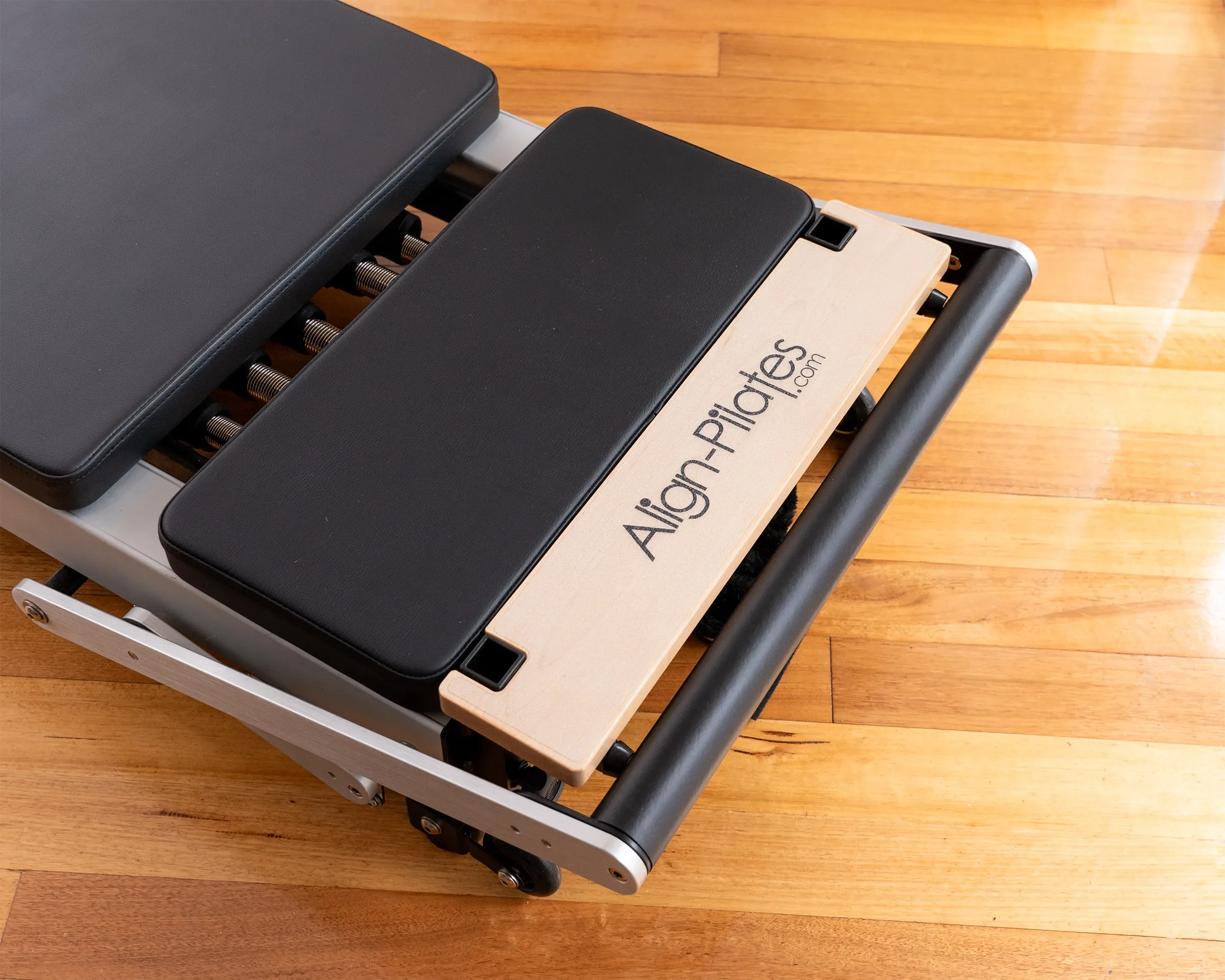 Close-up of an Align-Pilates reformer showing the wooden footbar, padded platform and springs on a polished studio floor.