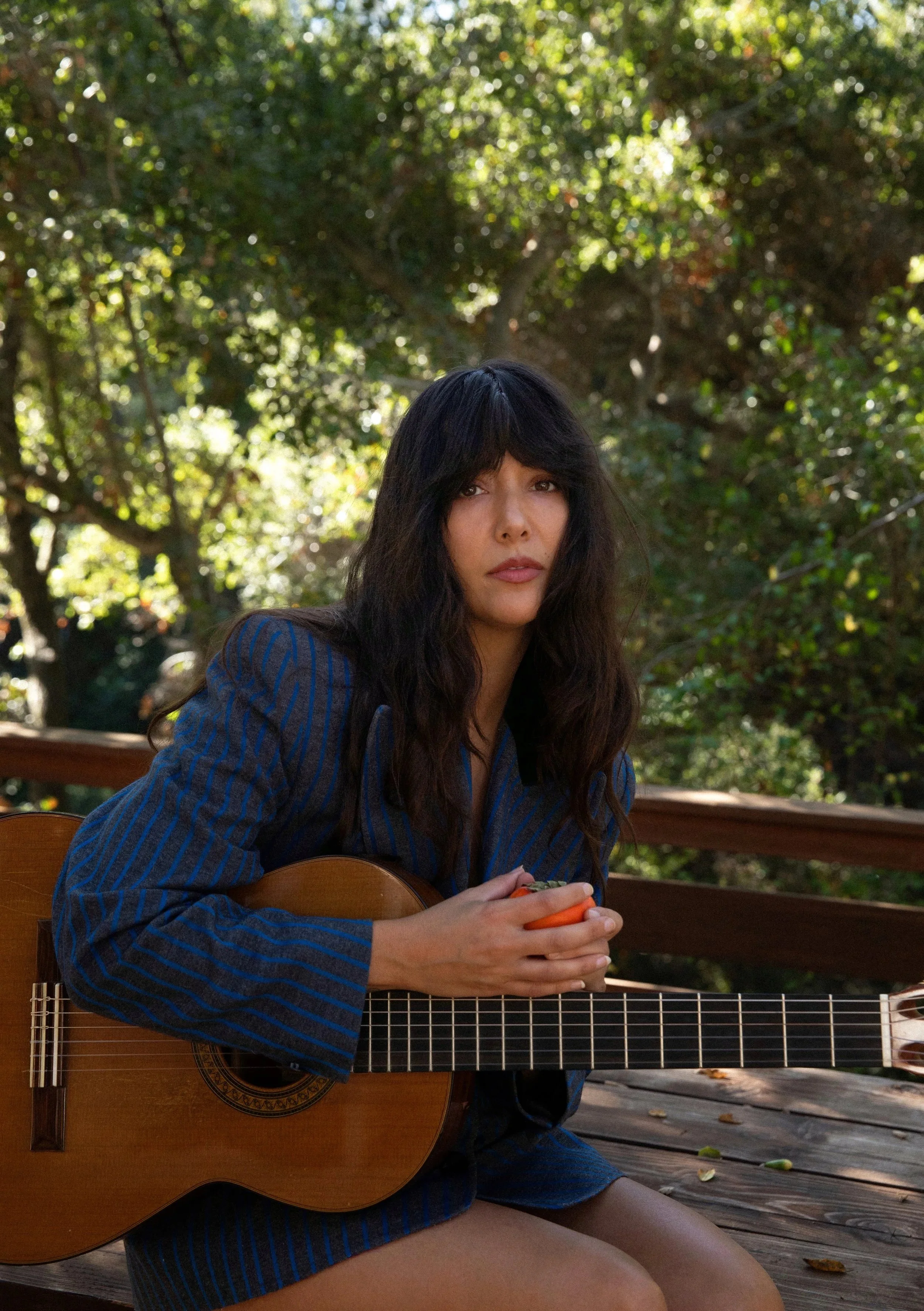 A woman with dark, wavy hair sitting outdoors on a wooden deck, holding an orange object, with a guitar resting on her lap, surrounded by green trees and foliage in the background.