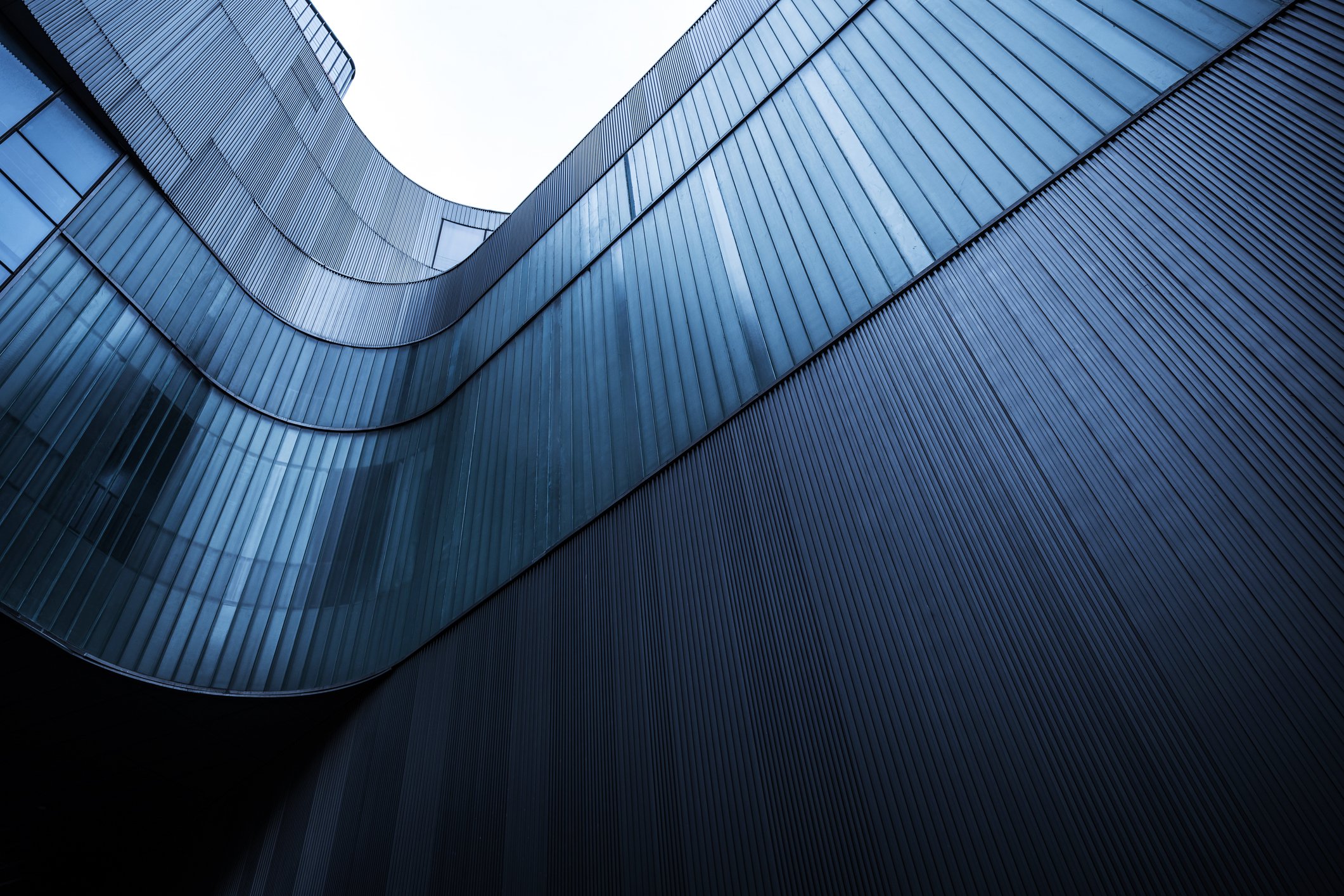 The image shows a modern glass building with curved blue-tinted windows and vertical metal siding, viewed from below with the sky visible at the top.