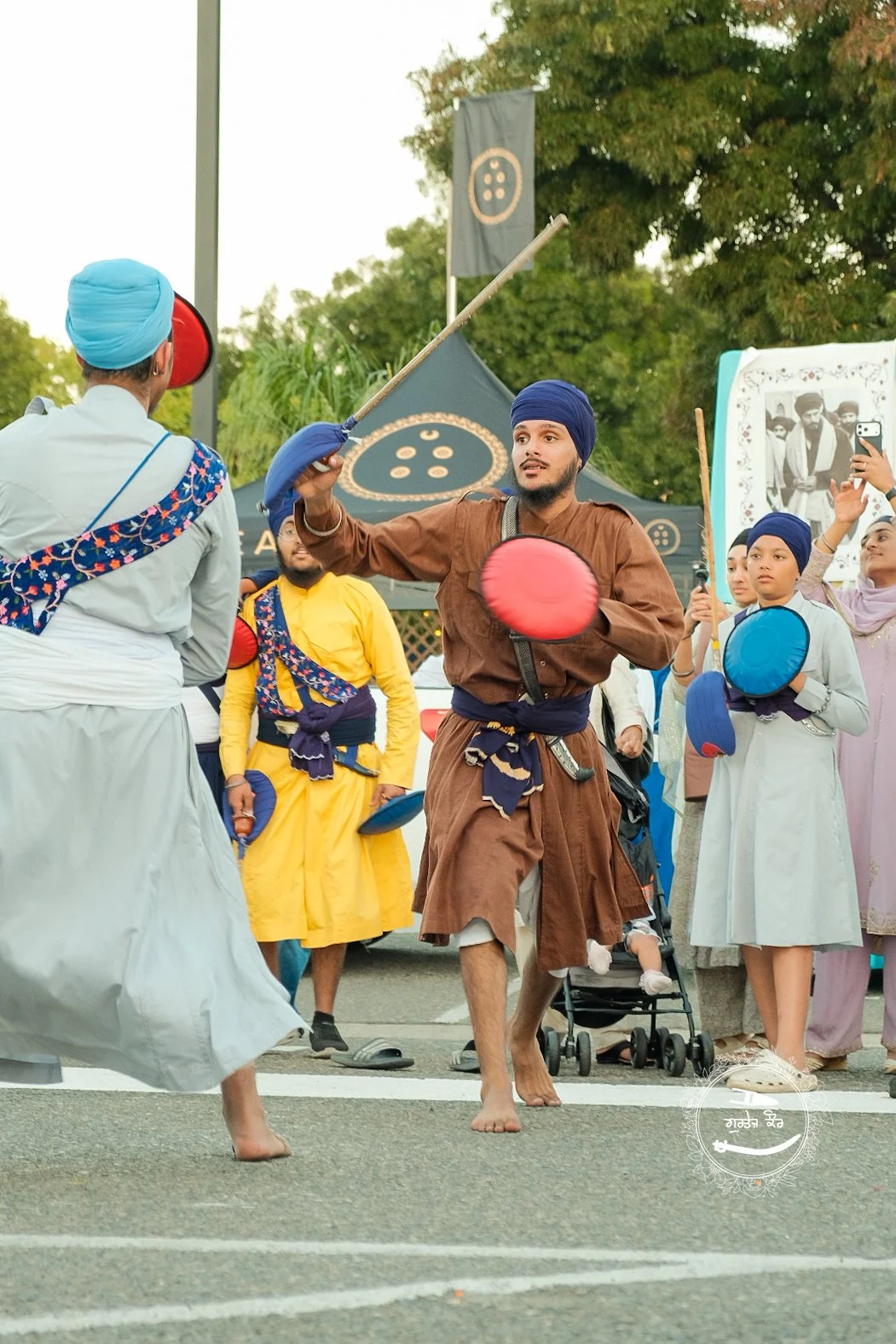 Sikh men and women playing traditional Gatka stick fighting during a cultural event outdoors, with tents and trees in the background, some people taking photos.