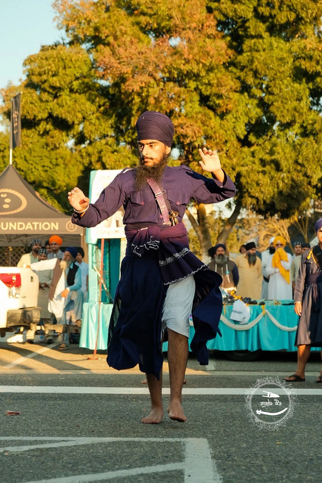 A man dressed in traditional Sikh attire performing a dance outdoors during a cultural event. He is barefoot, wearing a purple turban and dark purple and white clothing. There are people in traditional attire and a tent in the background, with large 