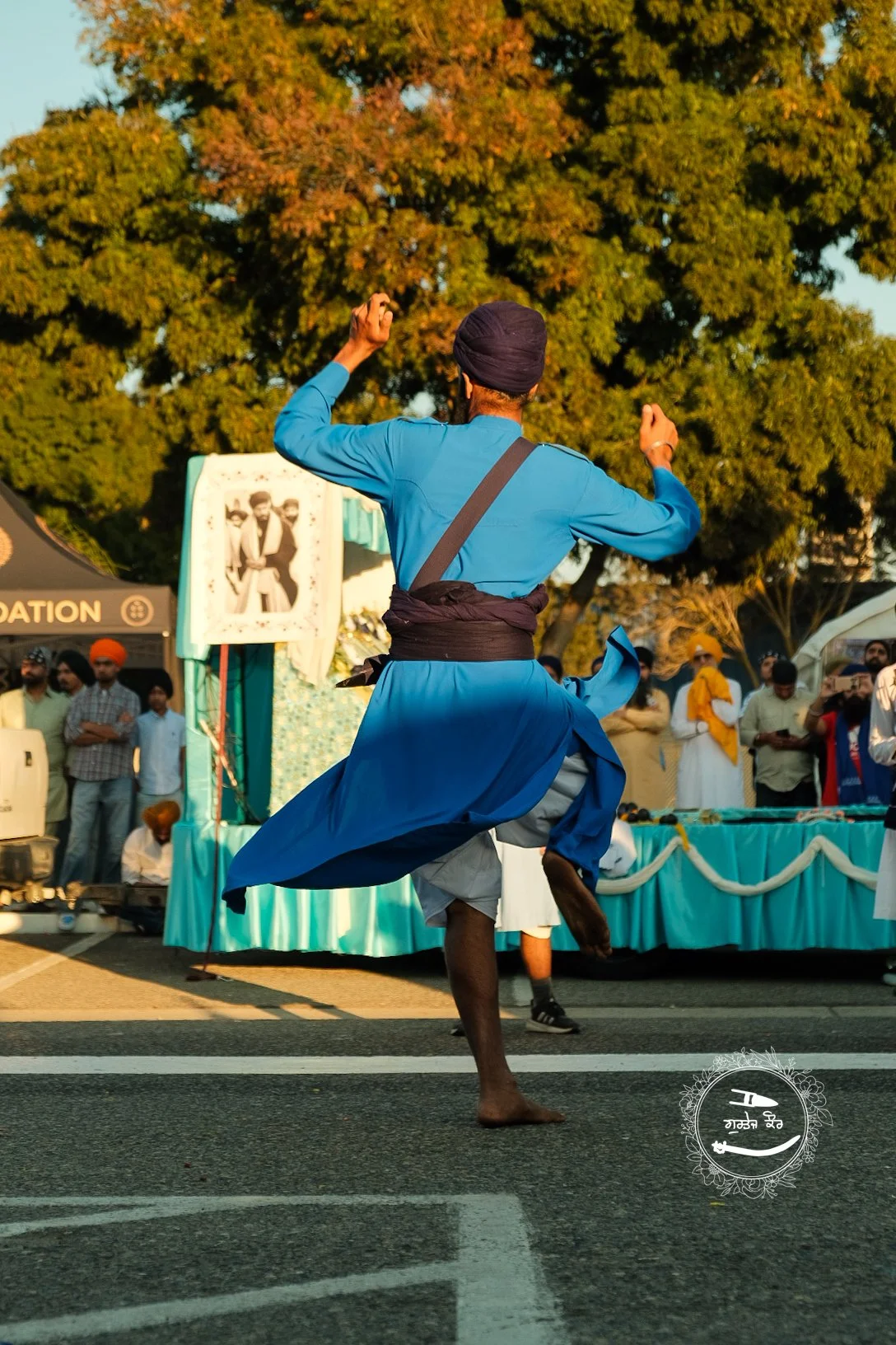 A man dressed in a blue traditional Punjabi outfit performing Bhangra dance on the street, with a crowd and decorated stage in the background, during a cultural festival.