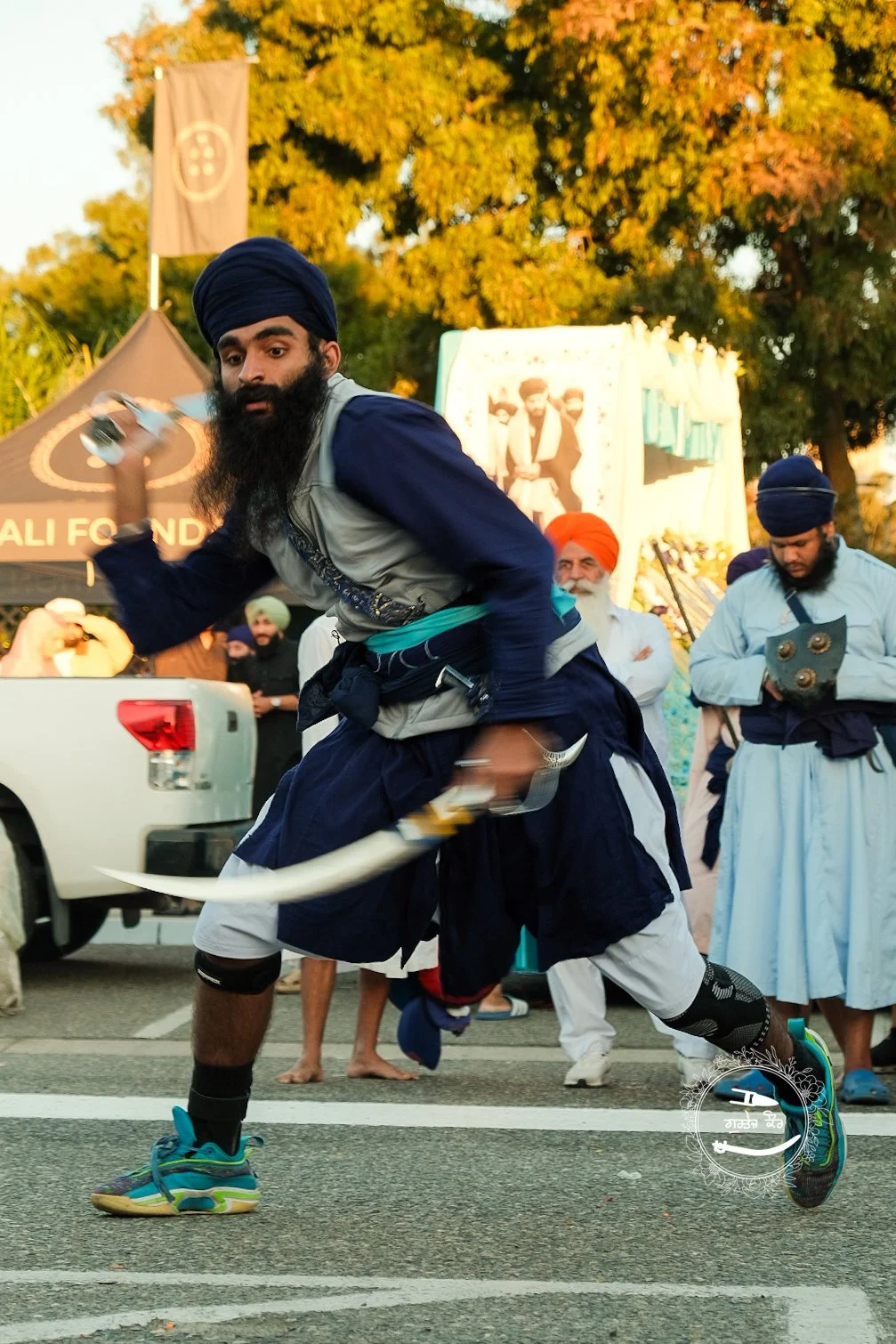A man wearing a turban and traditional Sikh attire participates in a cultural festival, performing an athletic move with a sword in hand. Several onlookers, including other men in traditional attire, watch in the background as tents and trees suggest